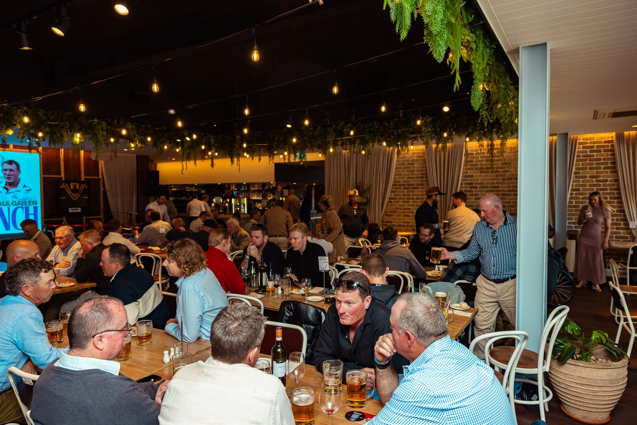 A crowded indoor restaurant or pub filled with people sitting at tables, enjoying drinks and conversations. The decor includes hanging string lights and green plants, with a brick wall and curtains in the background.