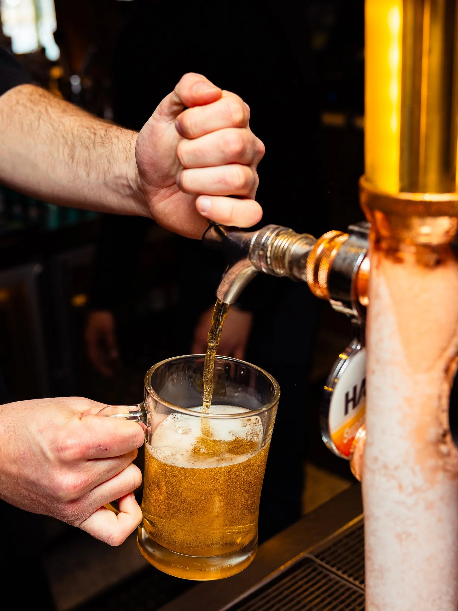 A person is pouring beer from a tap into a clear glass mug, which is held by another person. The beer is golden and foamy at the top, and the setting appears to be a bar or pub.