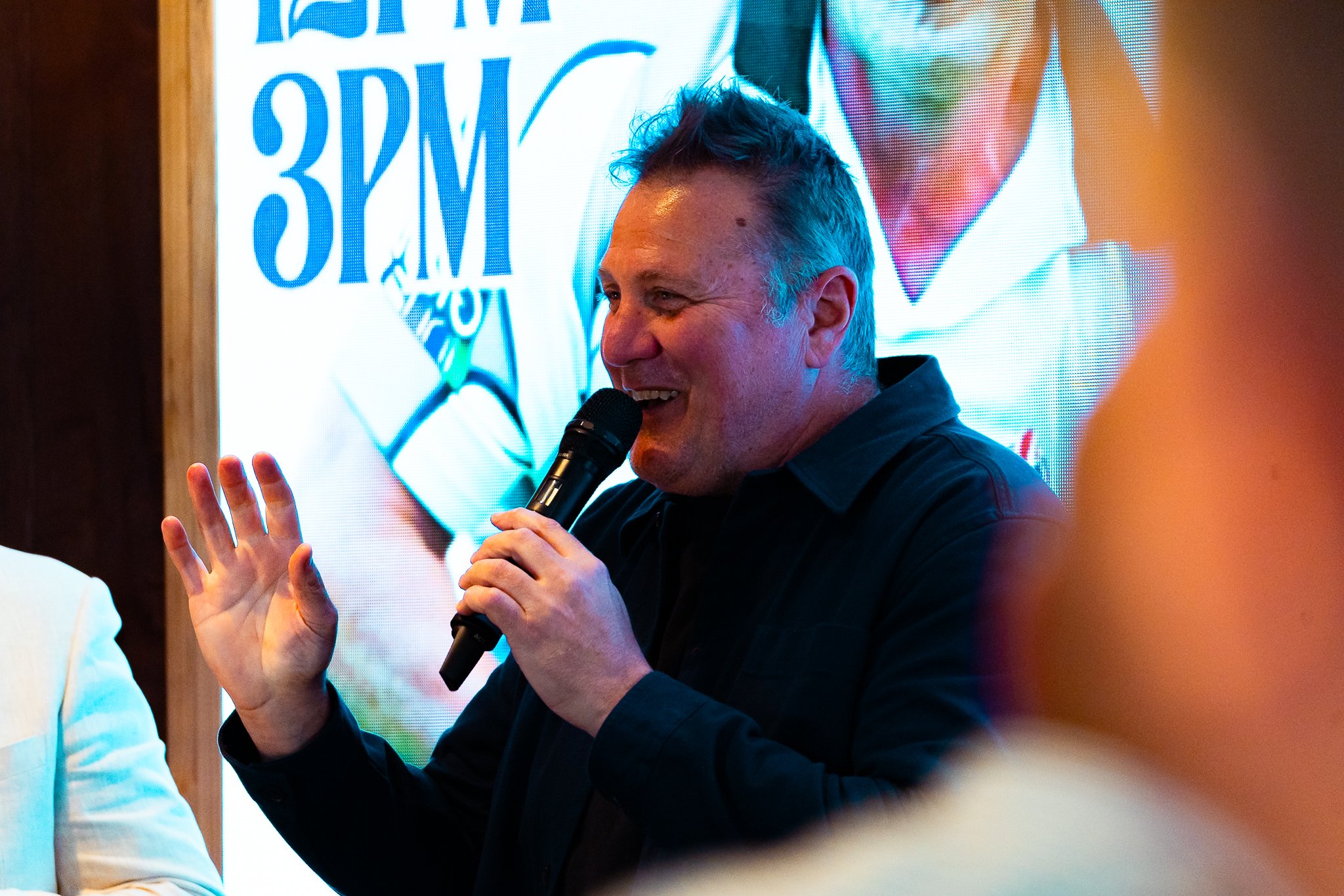 A man holding a microphone and speaking while gesturing with his hand during an event. A large screen behind him displays text and images, and there are some blurred people in the foreground.