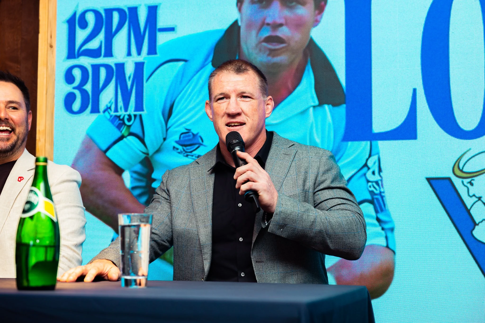 A man in a gray blazer and black shirt speaking into a microphone at a panel discussion. Behind him, there is a large screen displaying a close-up of his face and some text and graphics. Part of another person in a white jacket is visible on the left