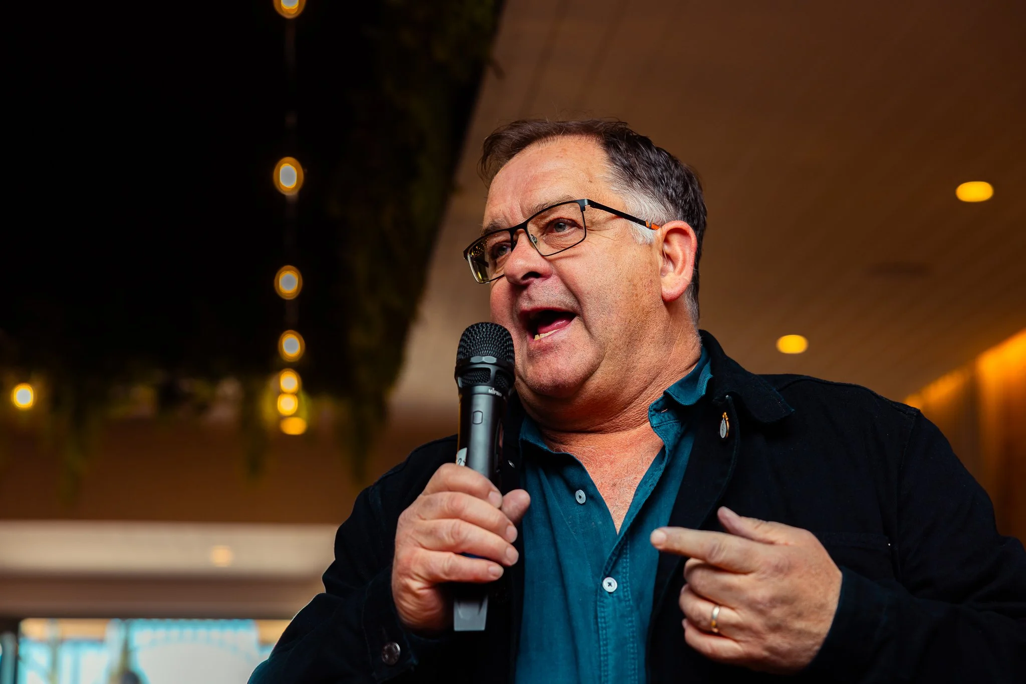 A man with glasses speaking into a microphone in an indoor setting with warm lighting.