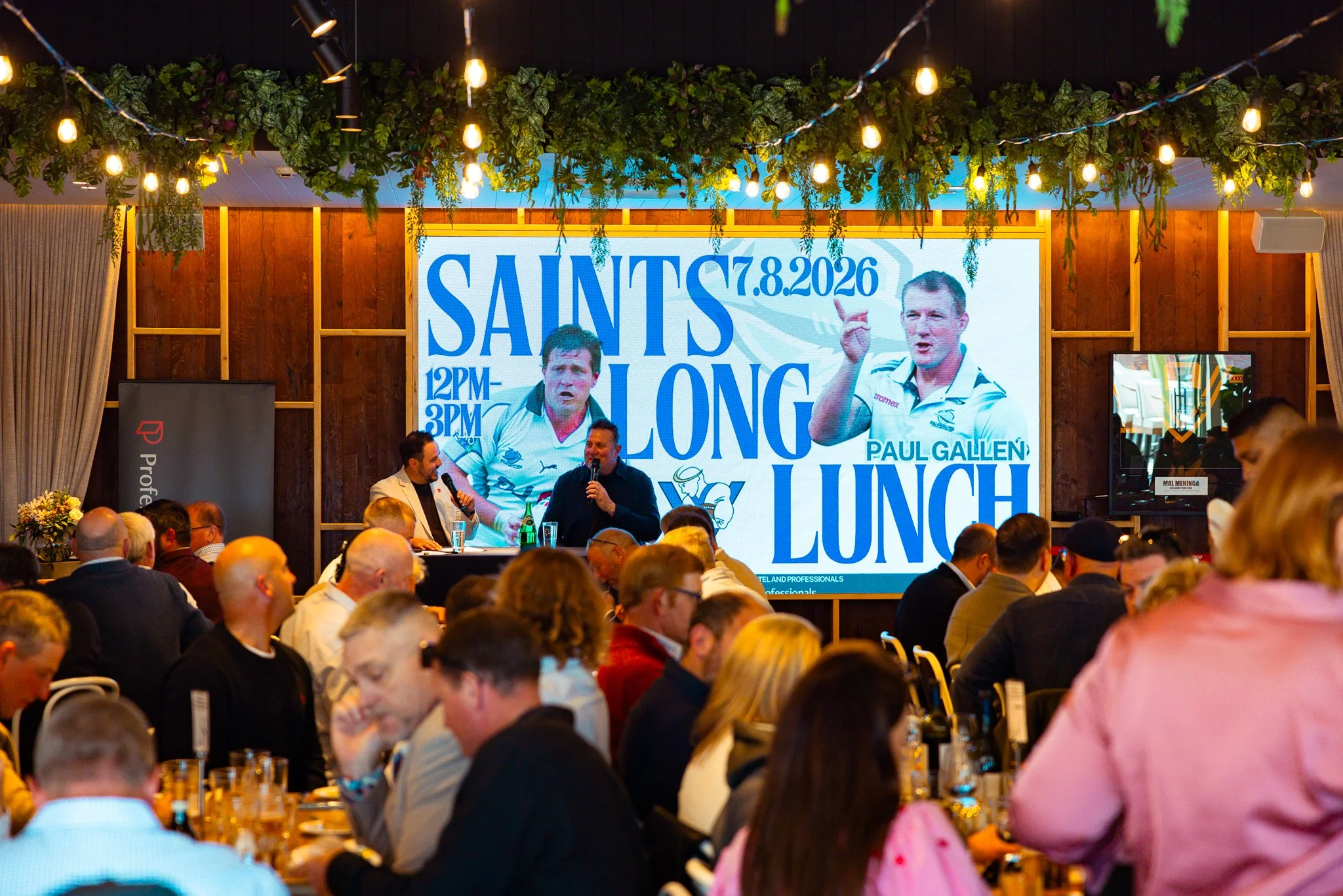 Large indoor event with a crowd of people seated at tables, facing a stage with a large digital screen showing a promotional poster for a lunch event featuring Paul Gallen on August 7, 2026. The poster displays images of Paul Gallen, one with him poi