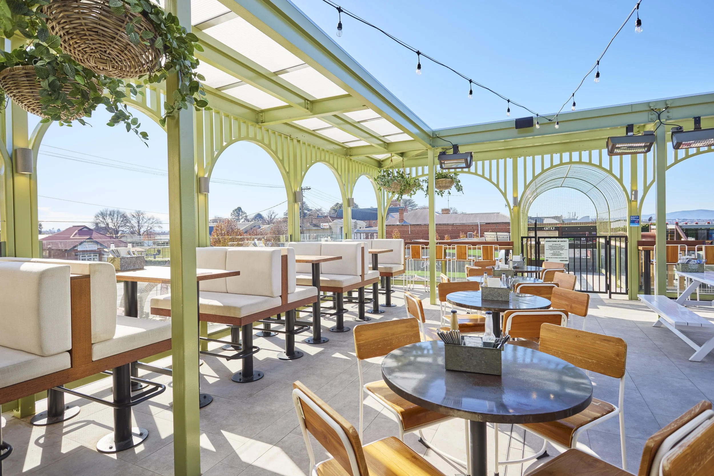 Outdoor patio area with wooden and cushioned seating, hanging plants, string lights, and a view of a neighborhood under a clear blue sky.