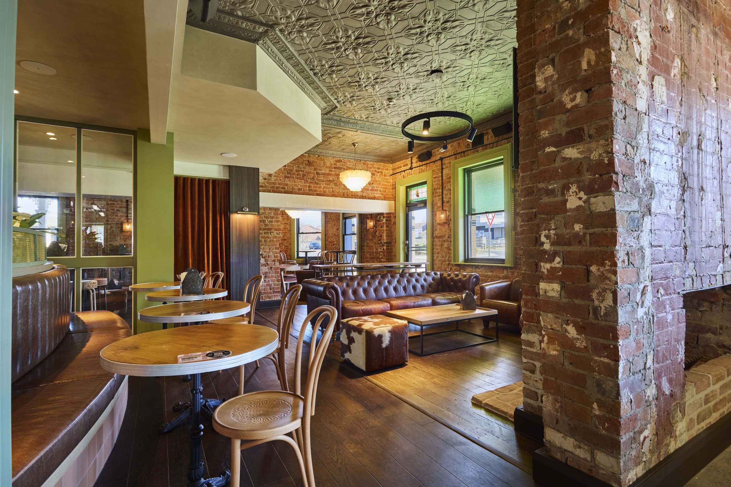 Interior of a cozy restaurant with exposed brick walls, decorative ceiling tiles, and wooden flooring. There are round tables with wooden chairs, leather tufted sofas, and a large window allowing natural light to brighten the space.