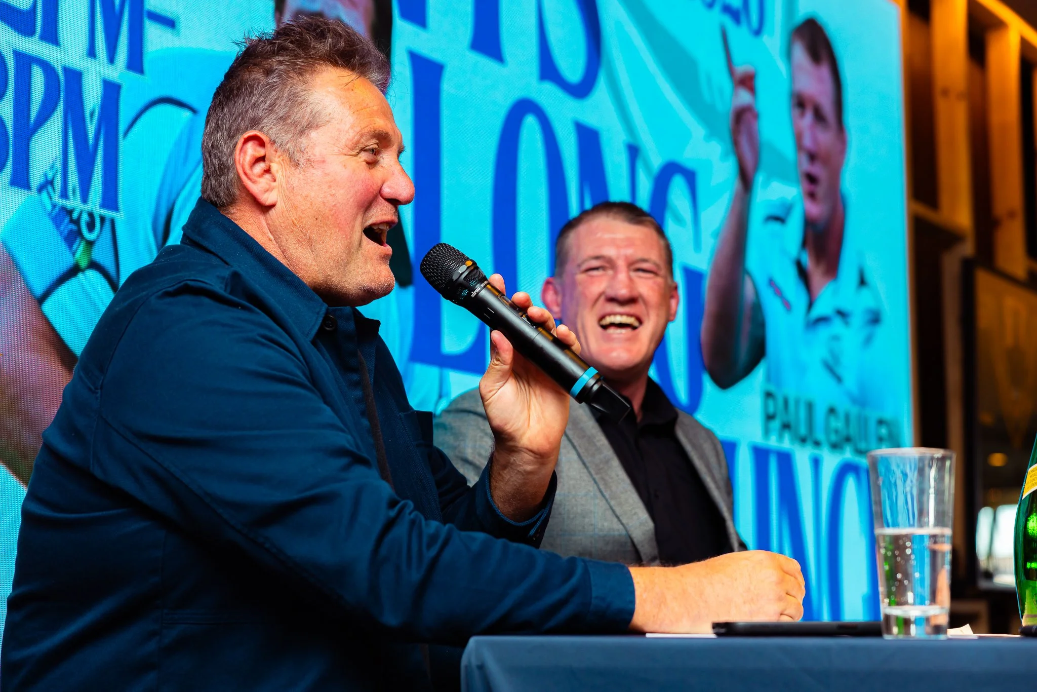 Two men sitting at a panel discussion or event. The man on the left is speaking into a microphone, while the man on the right is smiling. There is a large screen behind them displaying images and text.