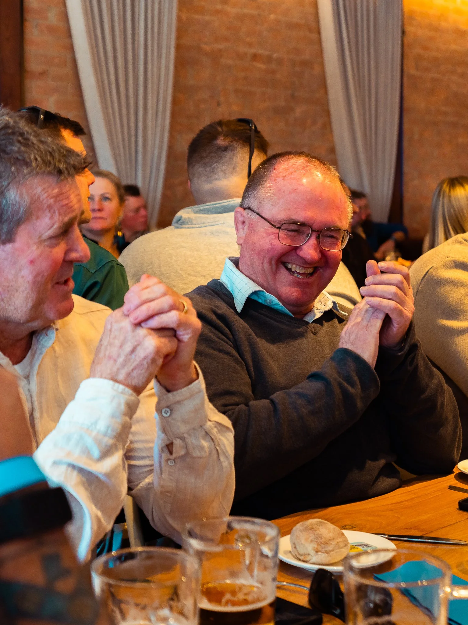 People sitting at a table in a restaurant, including a smiling man wearing glasses and a gray sweater, and a woman in a beige shirt. There are glasses, a bread roll on a plate, and some napkins on the table. The background shows brick walls and curta