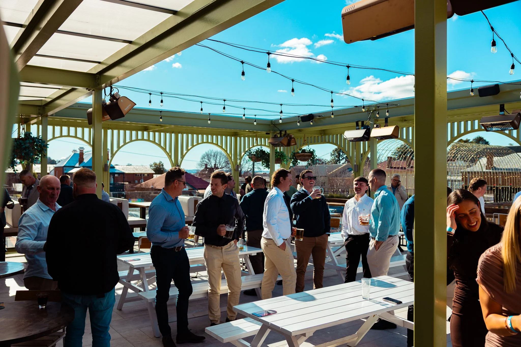 People socializing at an outdoor patio under a green pergola with string lights, blue sky, and buildings in the background.