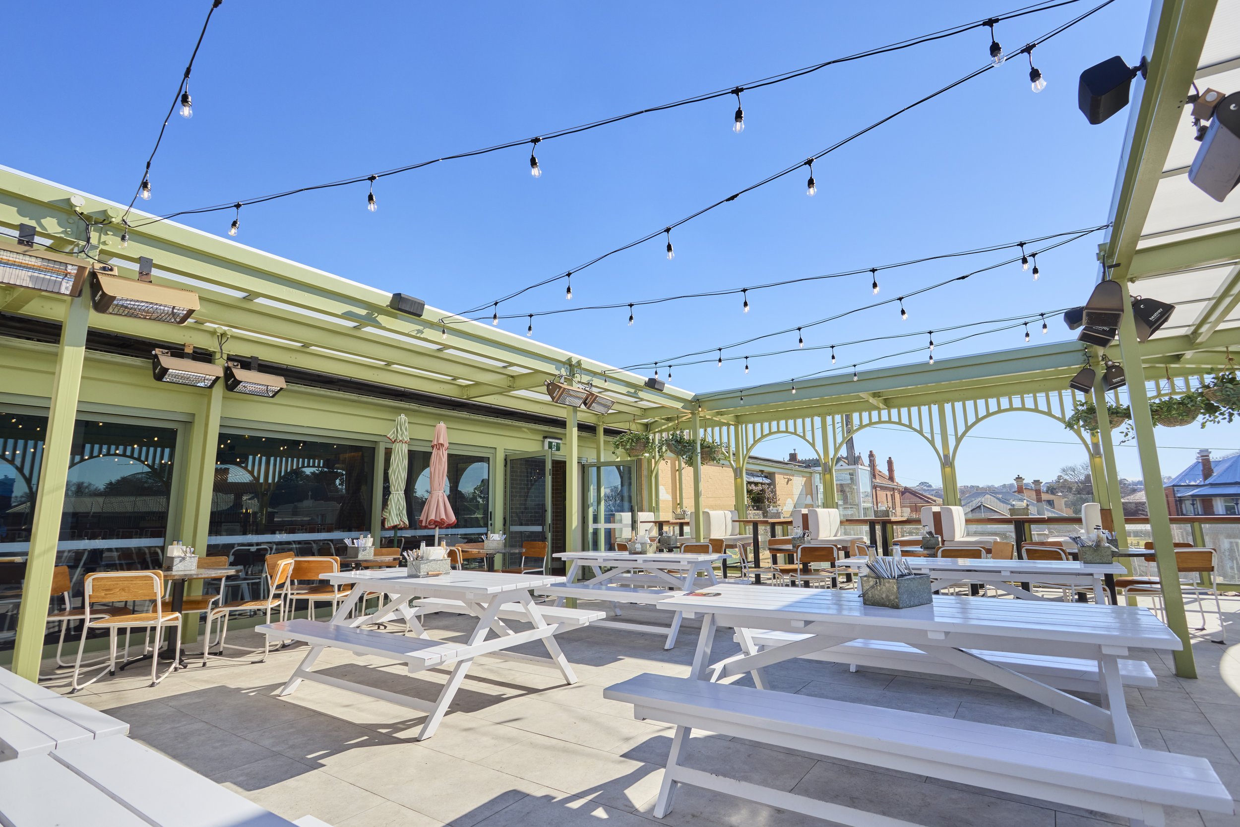 An outdoor patio area with white picnic tables and chairs, surrounded by a green pergola. String lights are hanging overhead, and there are heaters mounted on the pergola. The sky is clear and blue.