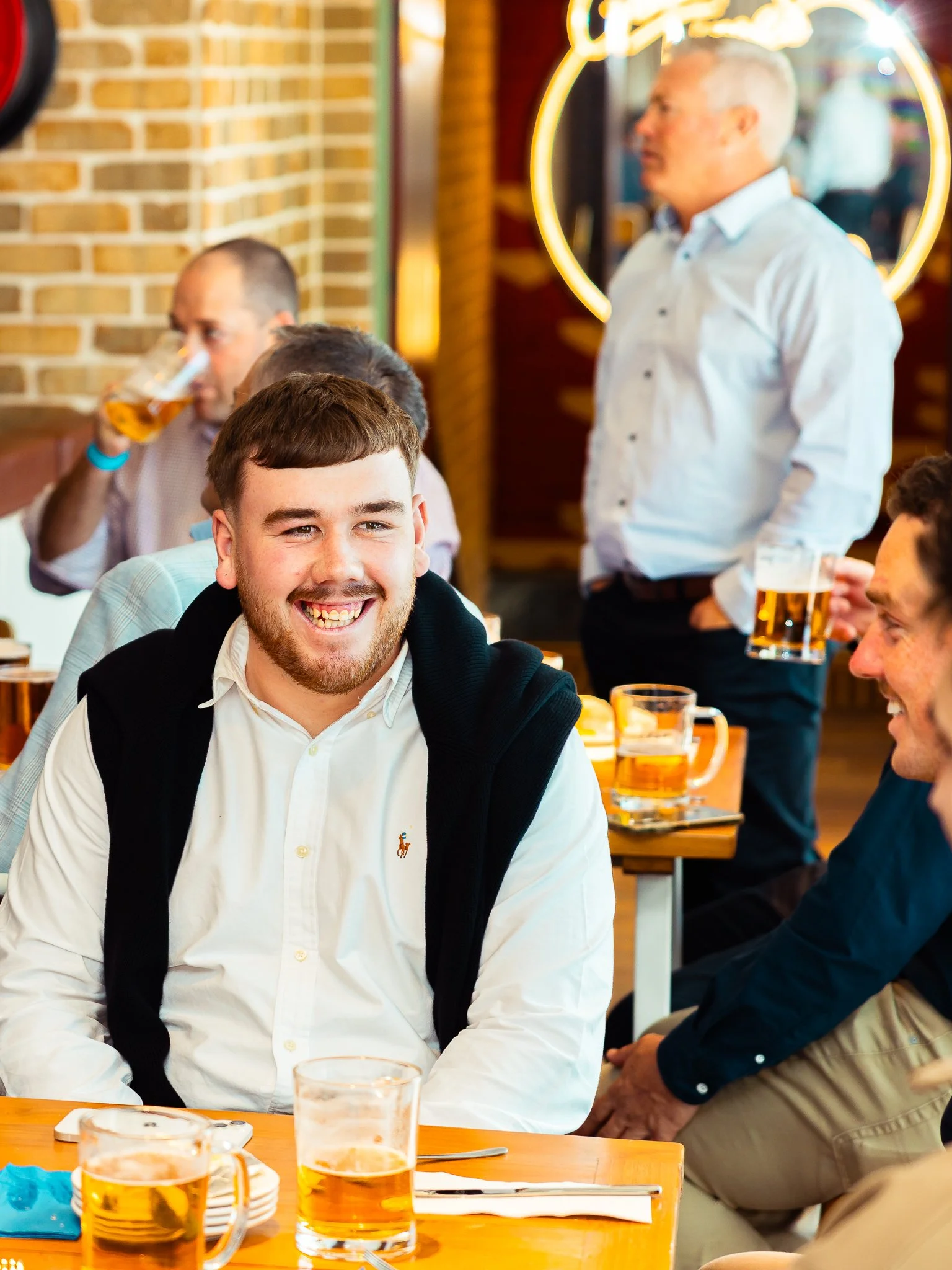 A group of people enjoying drinks at a restaurant or pub, with a young man smiling in the foreground, and others socializing in the background.