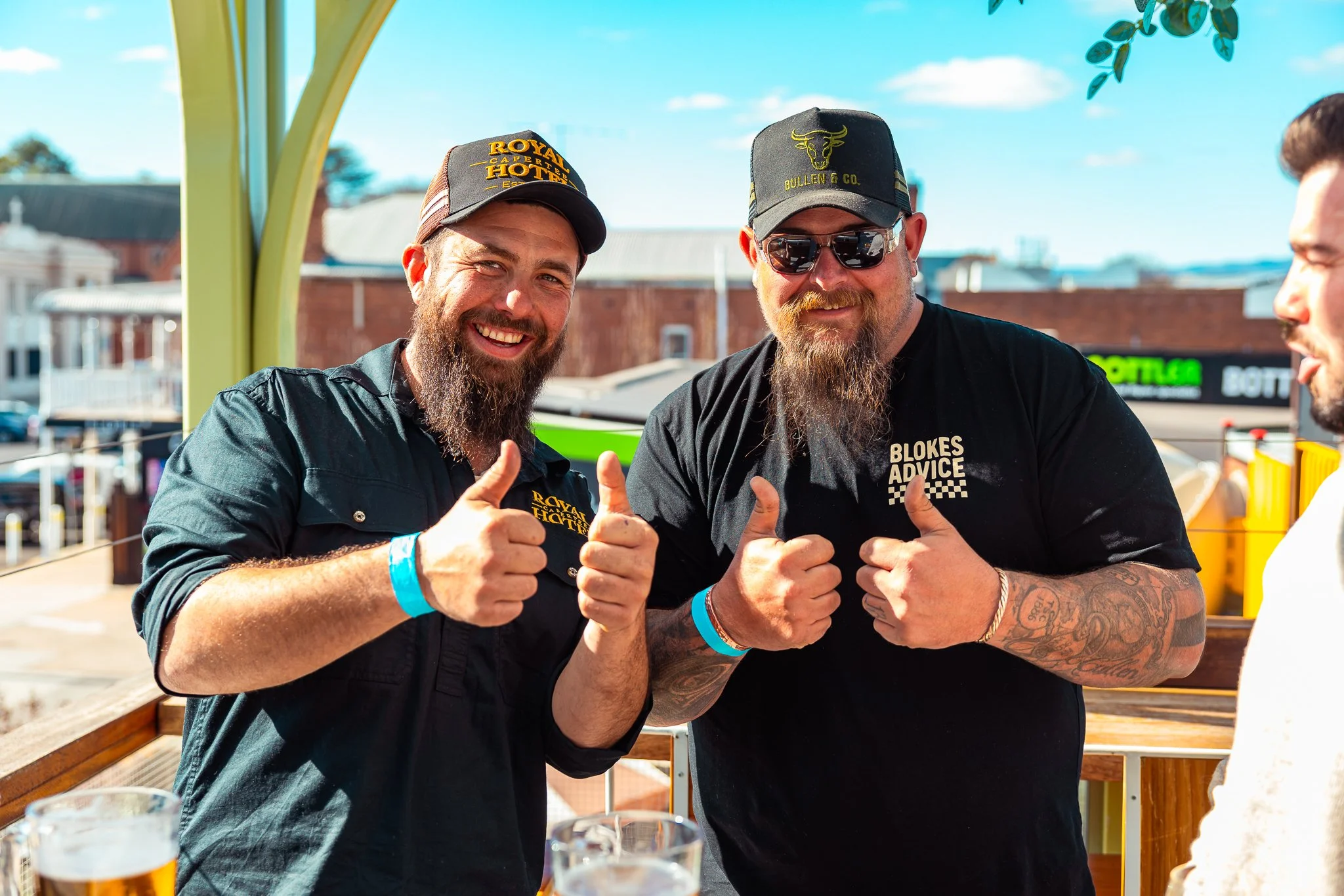 Two smiling men with beards showing thumbs up at an outdoor event, wearing black shirts and caps, with beverages in glasses in front of them.
