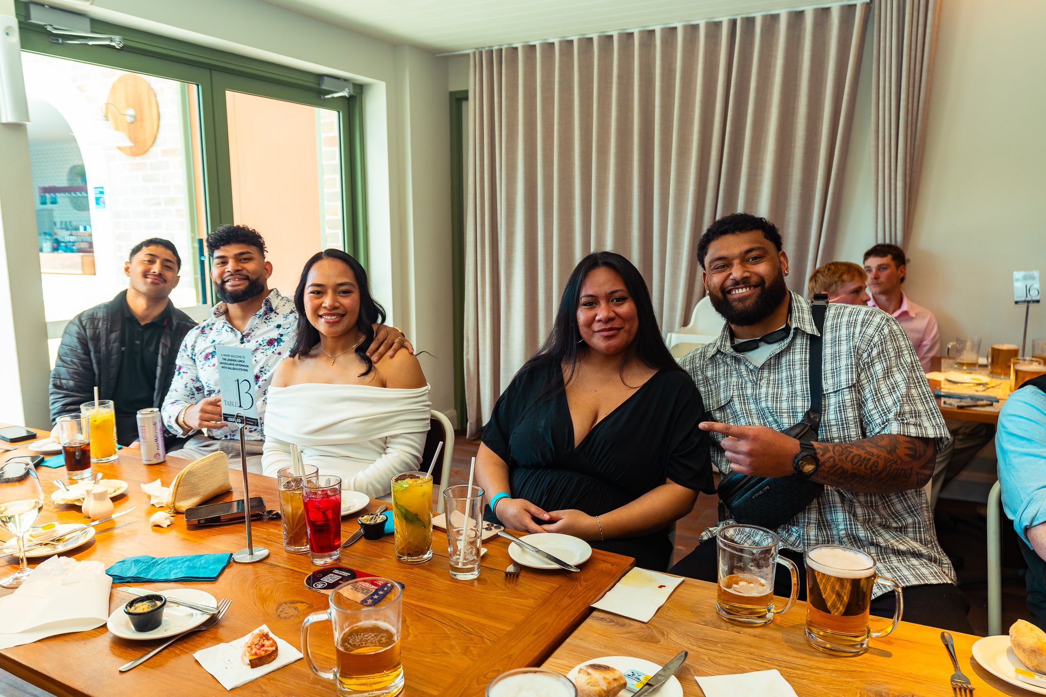 Group of six diverse people smiling and posing for a photo at a restaurant or cafe table with drinks, food, and tableware during a social gathering.