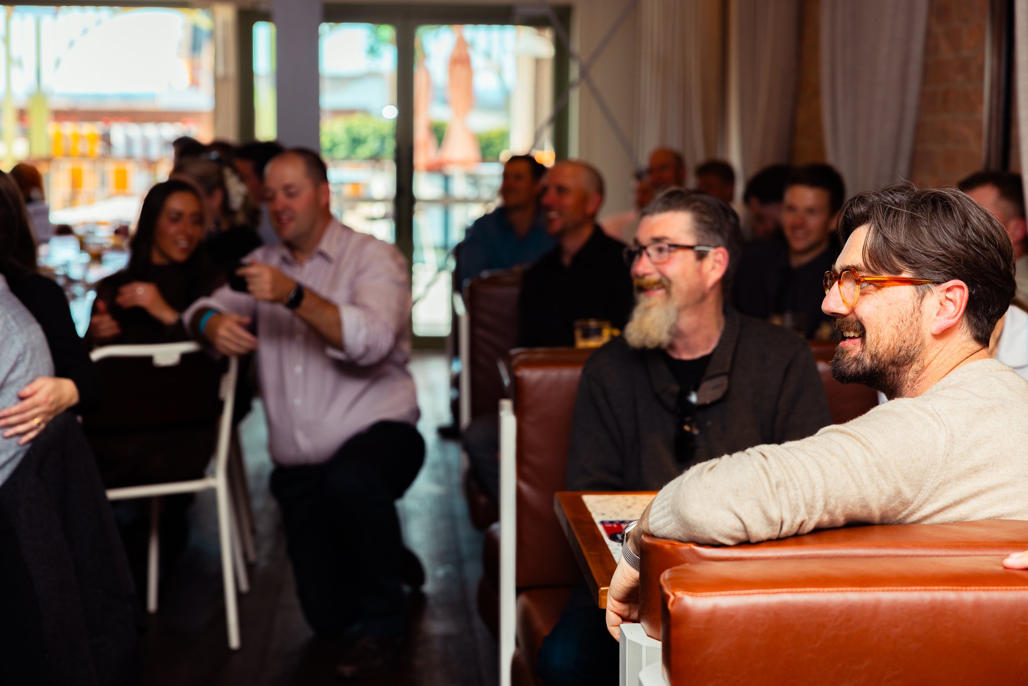 People gathered in a restaurant or café, smiling and engaged in conversation. The setting appears casual and cheerful, with large windows letting in natural light.
