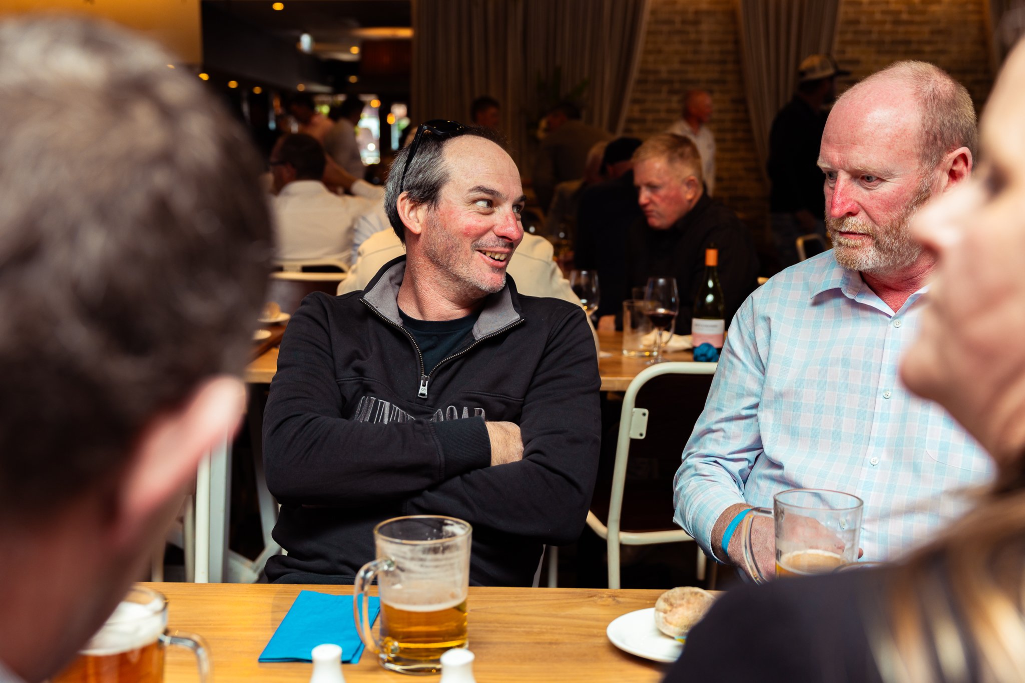 People socializing at a restaurant, with two men in focus smiling at each other across the table. Drinks and plates of food are visible on the table.