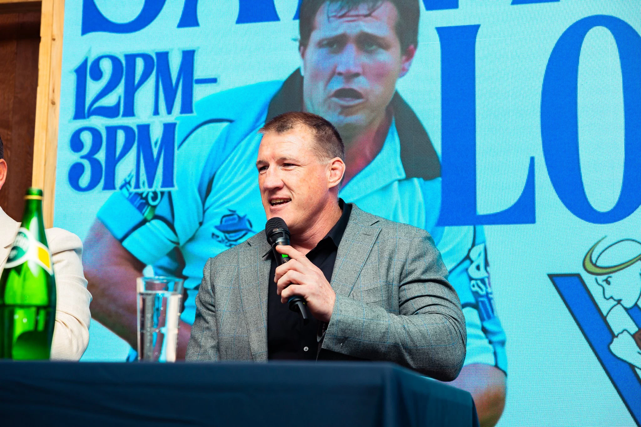A man in a gray checkered suit speaking into a microphone at a press conference, with a large digital screen behind him displaying a close-up image of his face and text referencing 12 p.m. and 3 p.m.