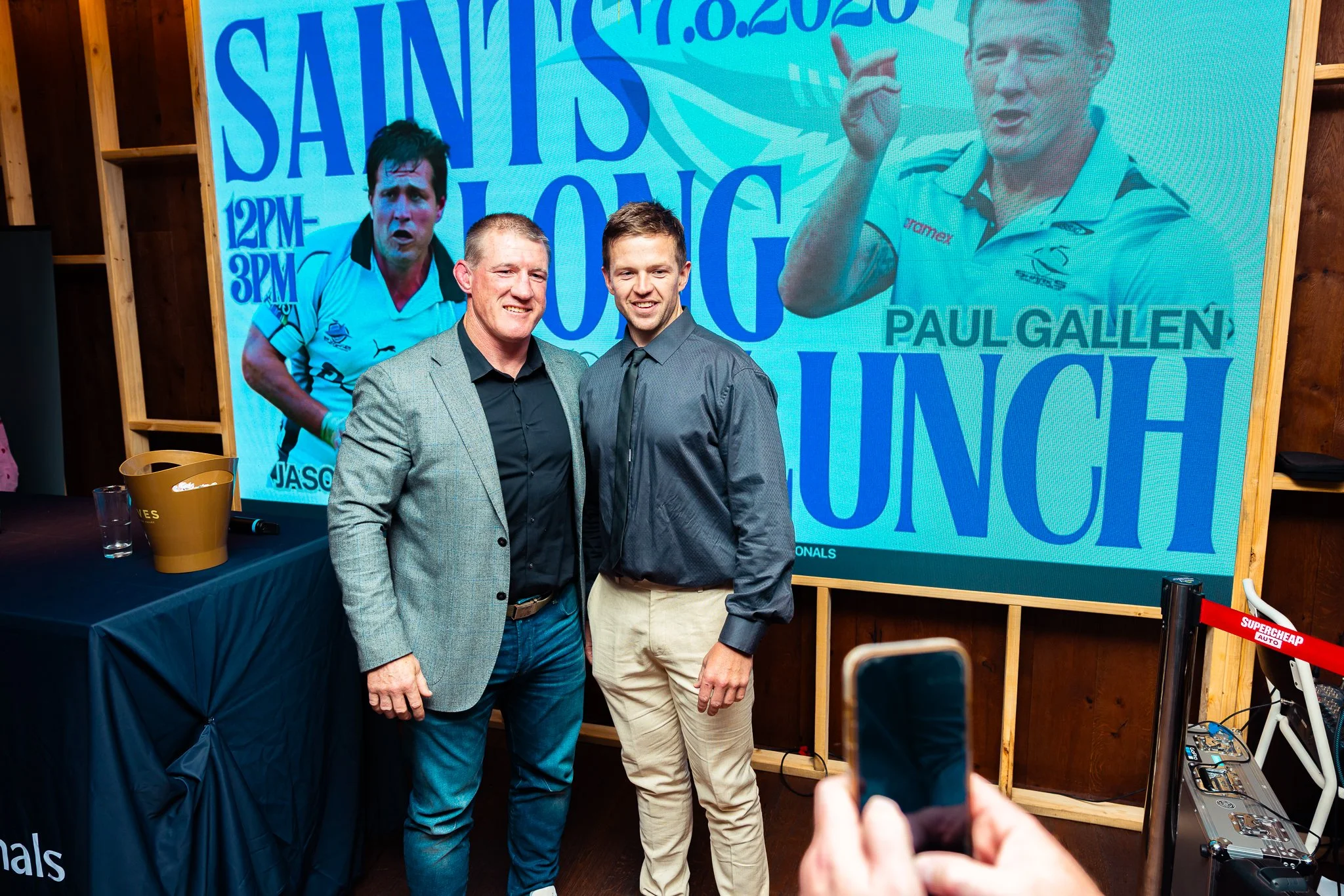 Two men smiling and dressed in business casual attire standing in front of a large banner featuring images of two athletes and event details at what appears to be a sports event or signing event.