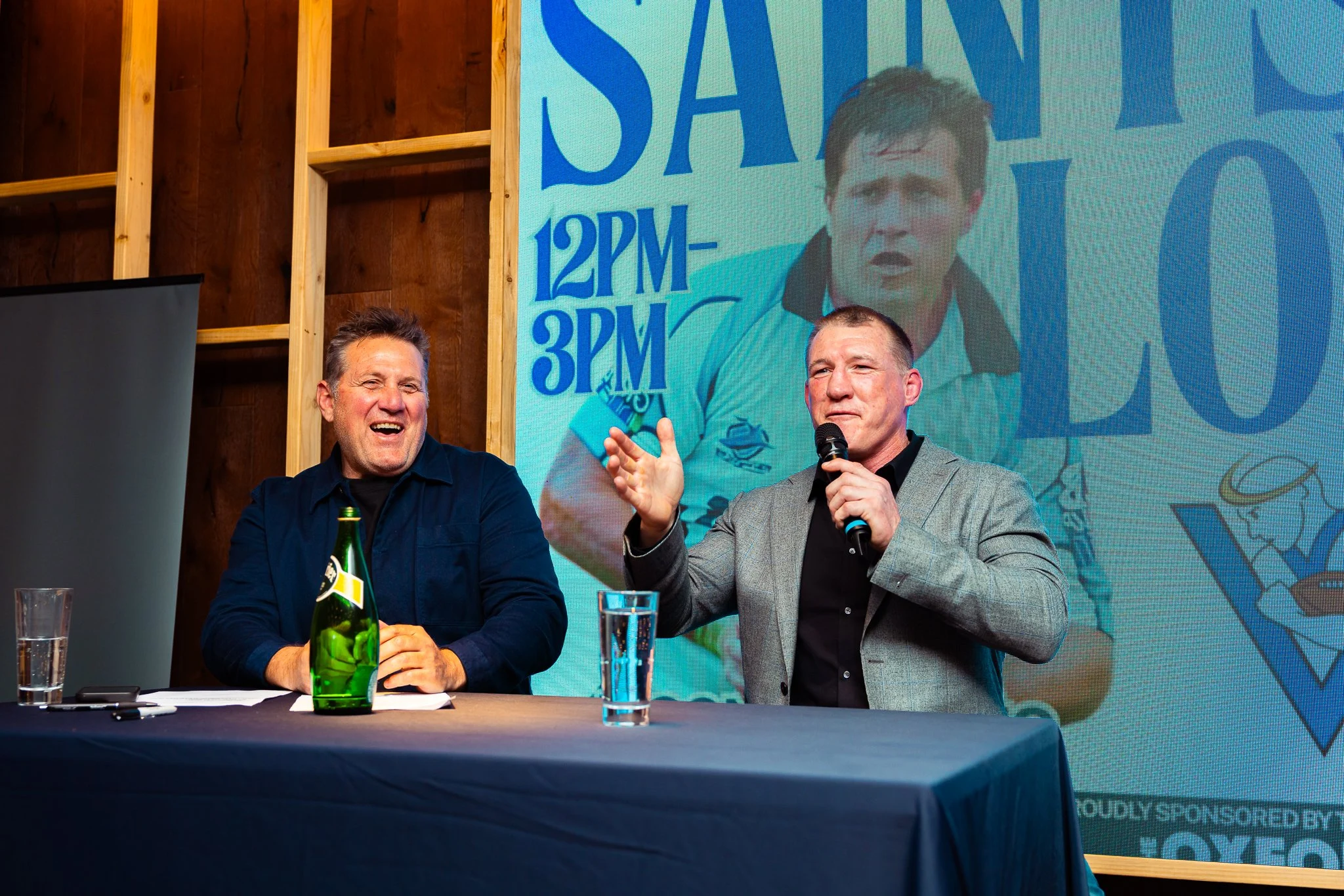 Two men sitting at a table during a discussion or interview. The man on the left has dark hair, an open mouth, and is smiling. The man on the right has short hair, is holding a microphone, and appears to be speaking. There are glasses of water and a 