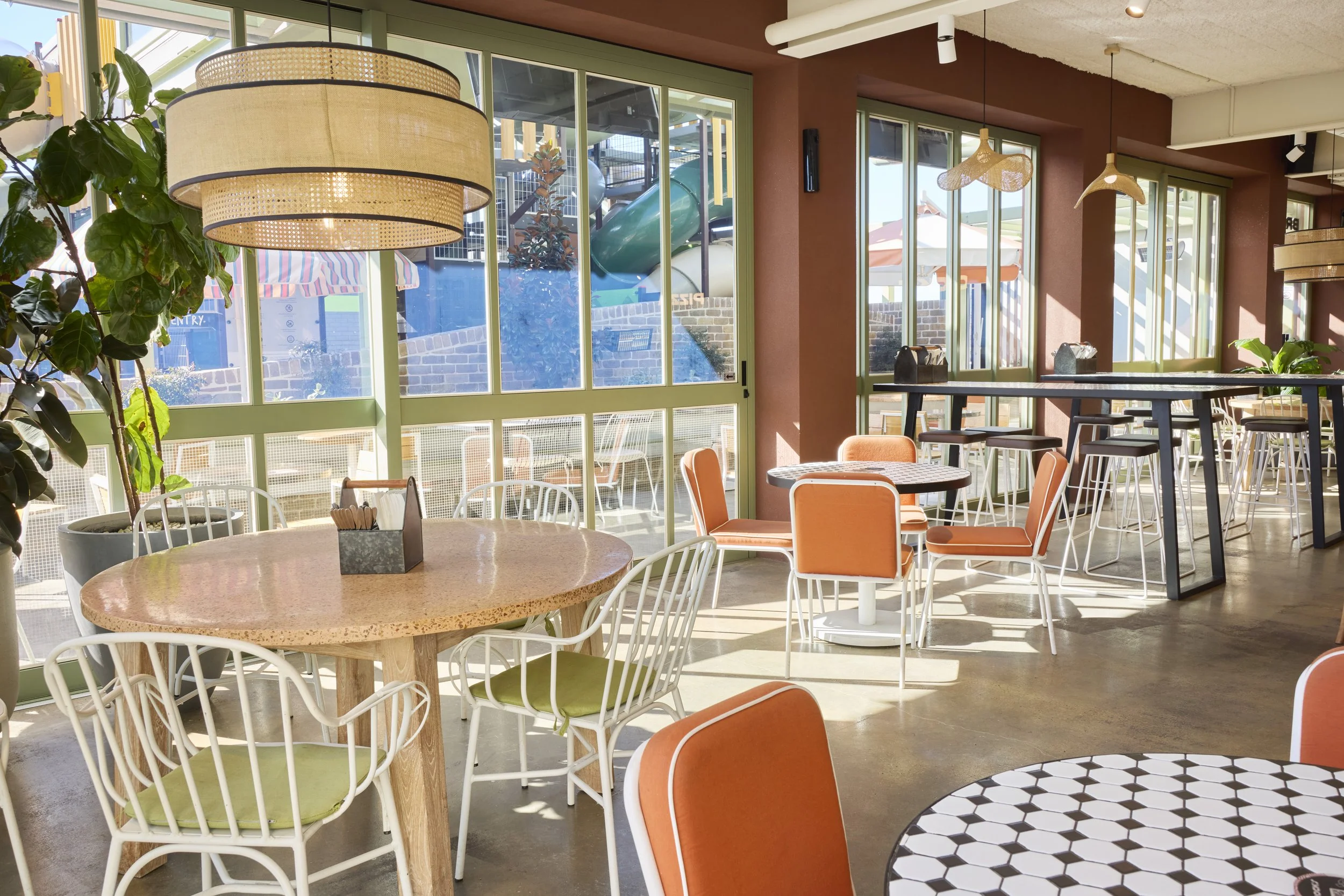 Inside a brightly lit cafe with large windows, orange and green chairs, white tables with black and white patterned tops, potted plants, and hanging pendant lights.