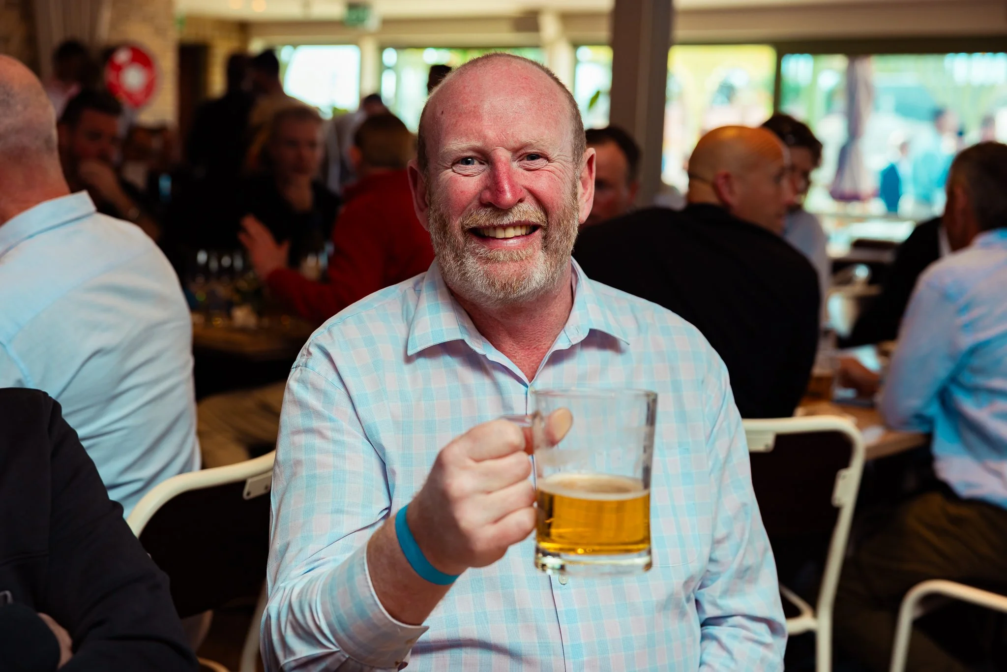 A smiling man with a beard holding a glass of beer in a crowded indoor event.