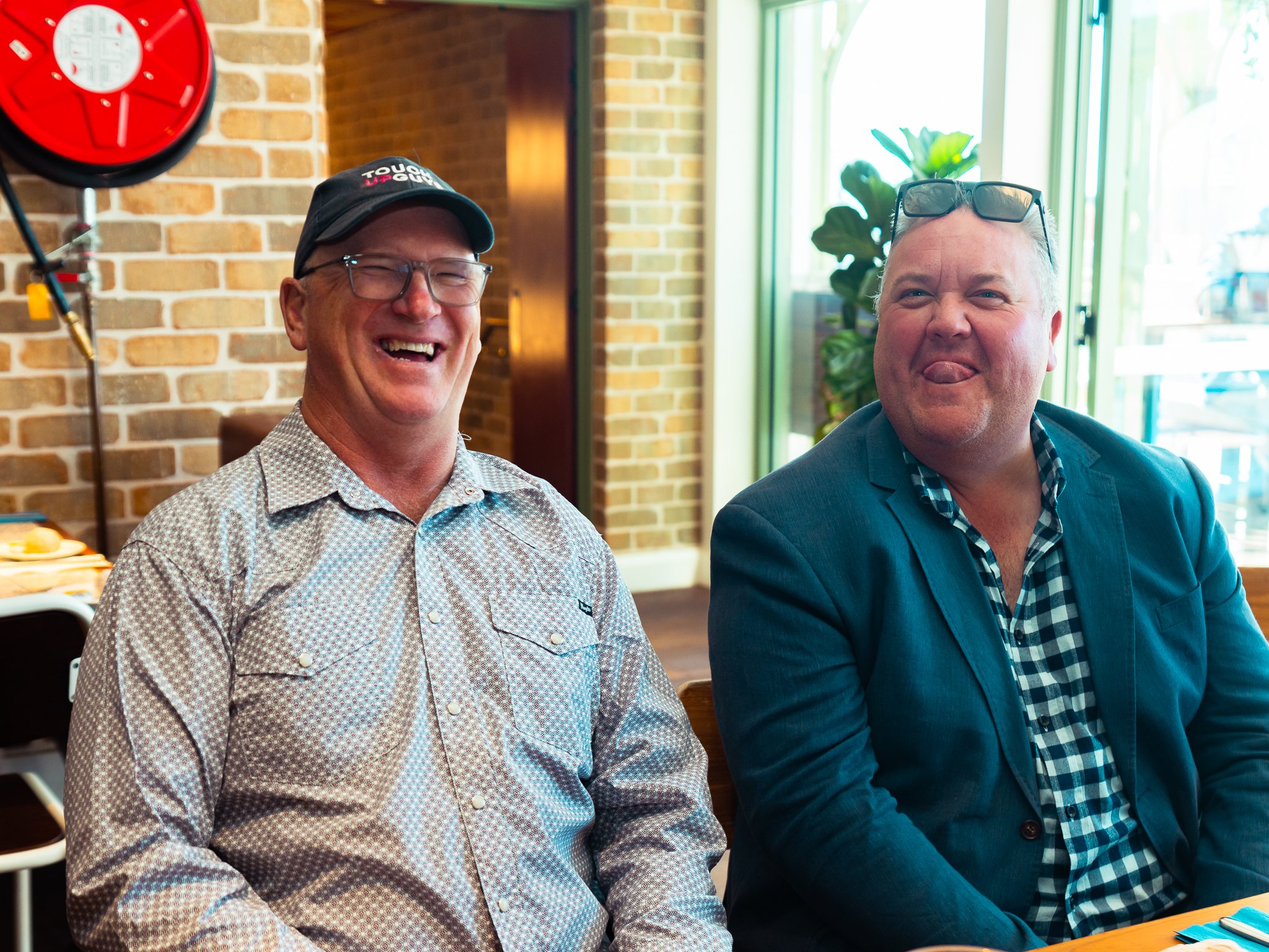 Two men sitting indoors, smiling and making funny faces, with brick walls and large windows in the background.