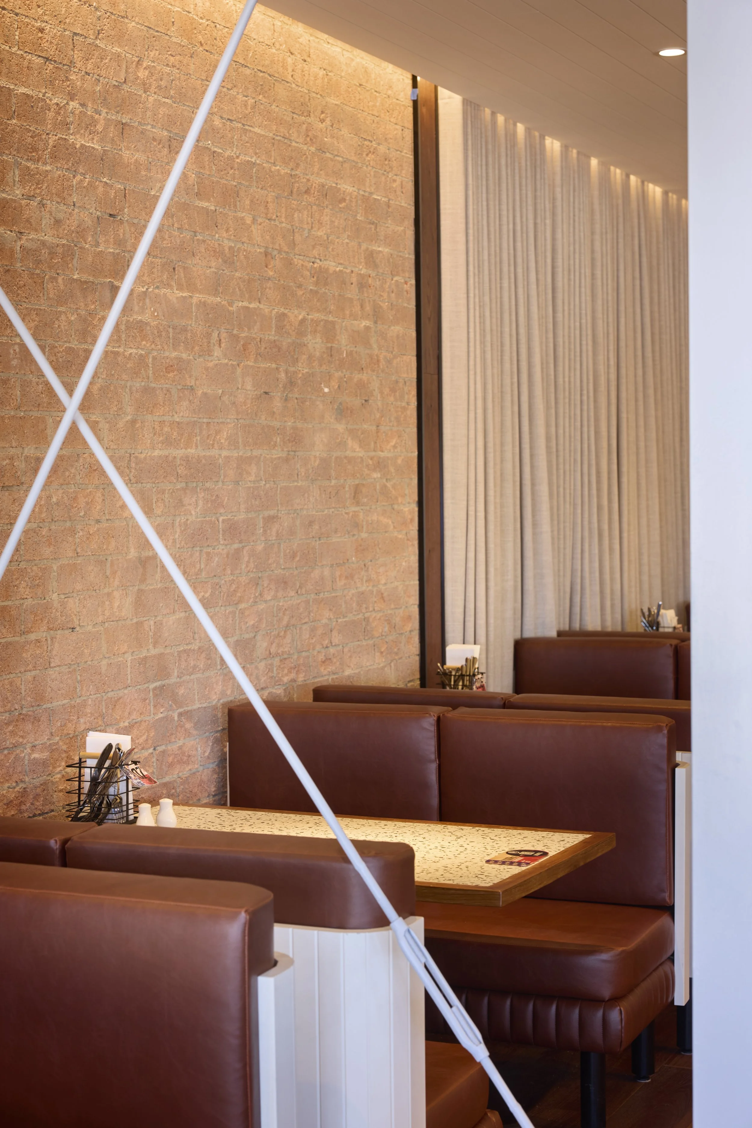 Interior of a restaurant with brown leather booth seating, a table with mustard color surface and cutlery, and a wall with exposed brick and floor-to-ceiling beige curtain.