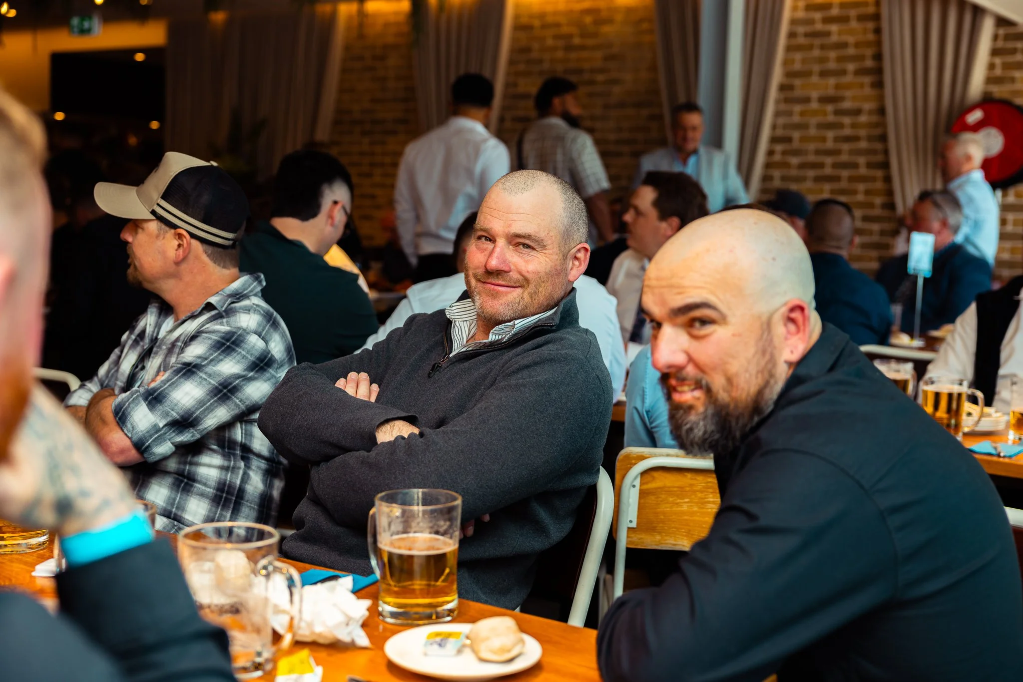 A group of men sitting at a table in a restaurant or pub, with some drinking beer. The man in the foreground has a shaved head and is smiling at the camera. The background shows other people standing and talking against a brick wall.