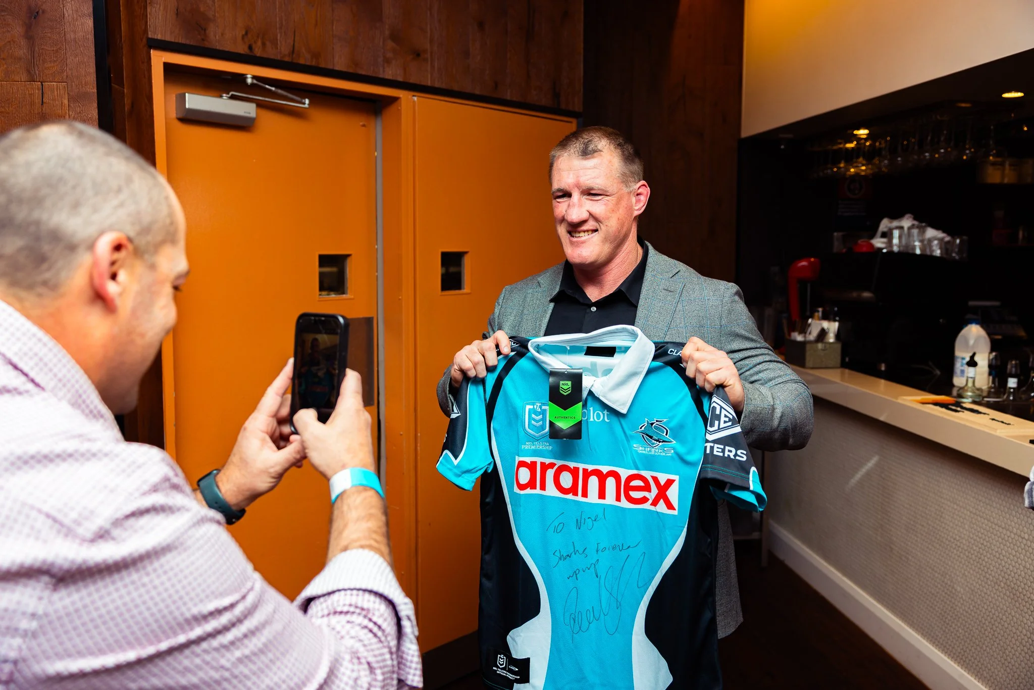 A man in a checkered shirt is taking a photo of another man in a sports jersey, who is smiling and holding the jersey in front of him. The jersey has sponsorship logos and a handwritten note.
