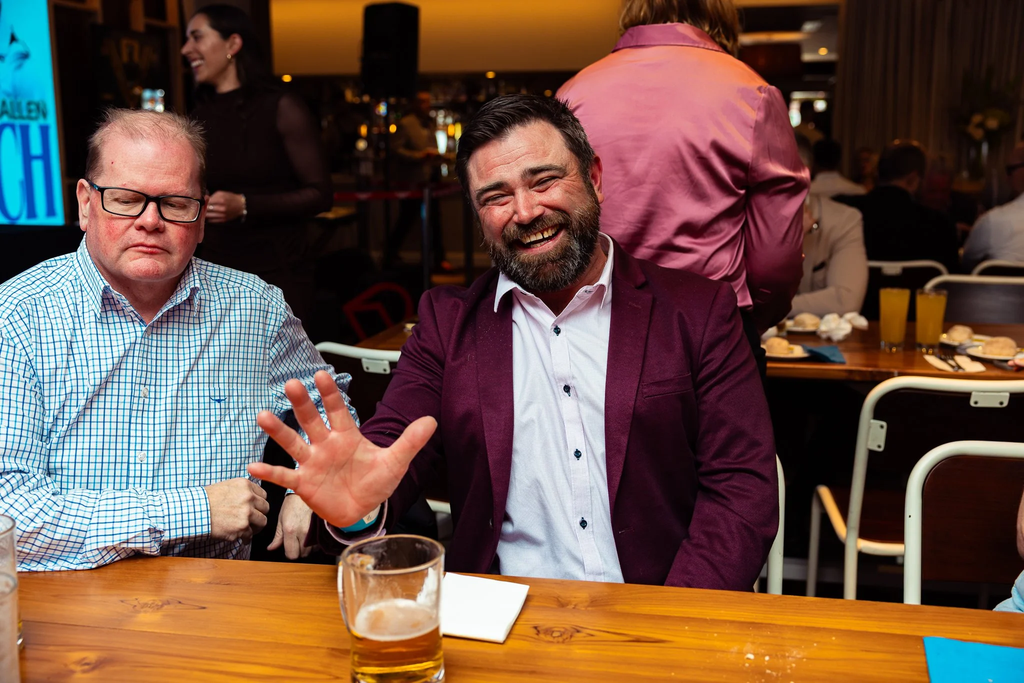 Two men sitting at a table in a restaurant or bar, one wearing glasses and a checkered shirt, looking down with a serious expression, the other with dark hair and a beard, wearing a maroon blazer and white shirt, smiling and waving at the camera. The