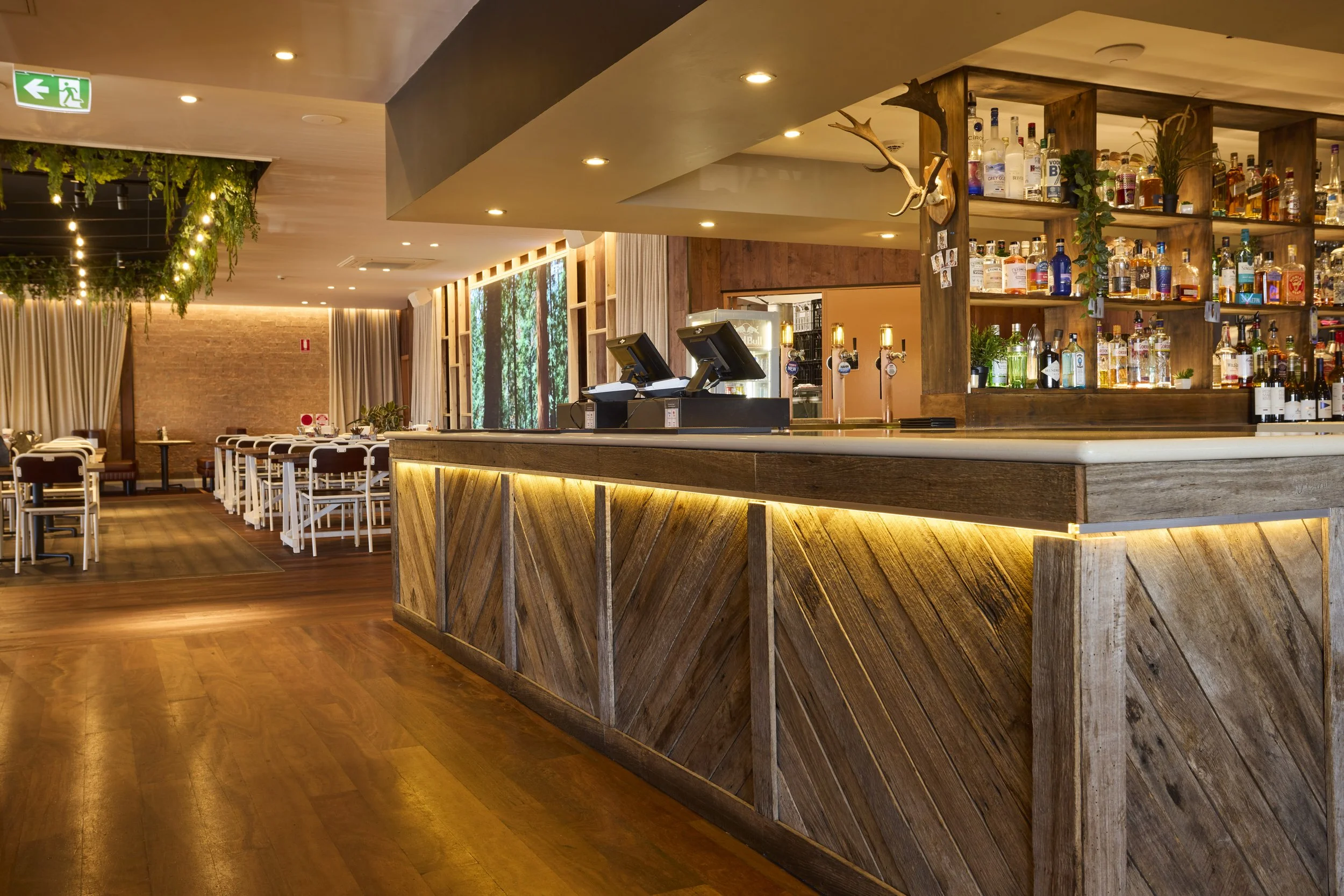 Interior of a modern bar with wooden decor, shelves stocked with liquor bottles, a bar counter with cash registers, and indoor plants hanging from the ceiling.