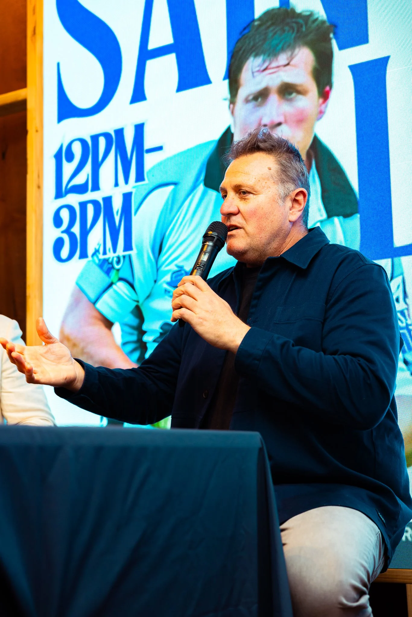 A man speaking into a microphone during a panel or presentation, with a large poster of a young man in the background displaying event details.