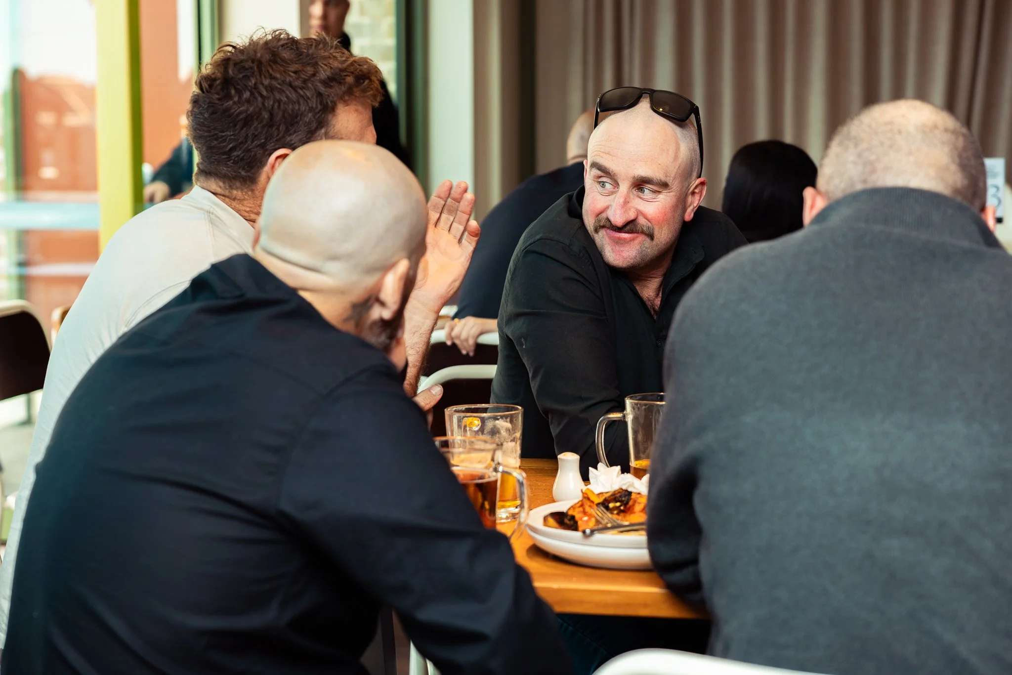 A group of men sitting around a wooden table in a restaurant, engaged in conversation. They have drinks on the table, and one man with a shaved head and sunglasses on his head is smiling and talking to another man with curly hair.