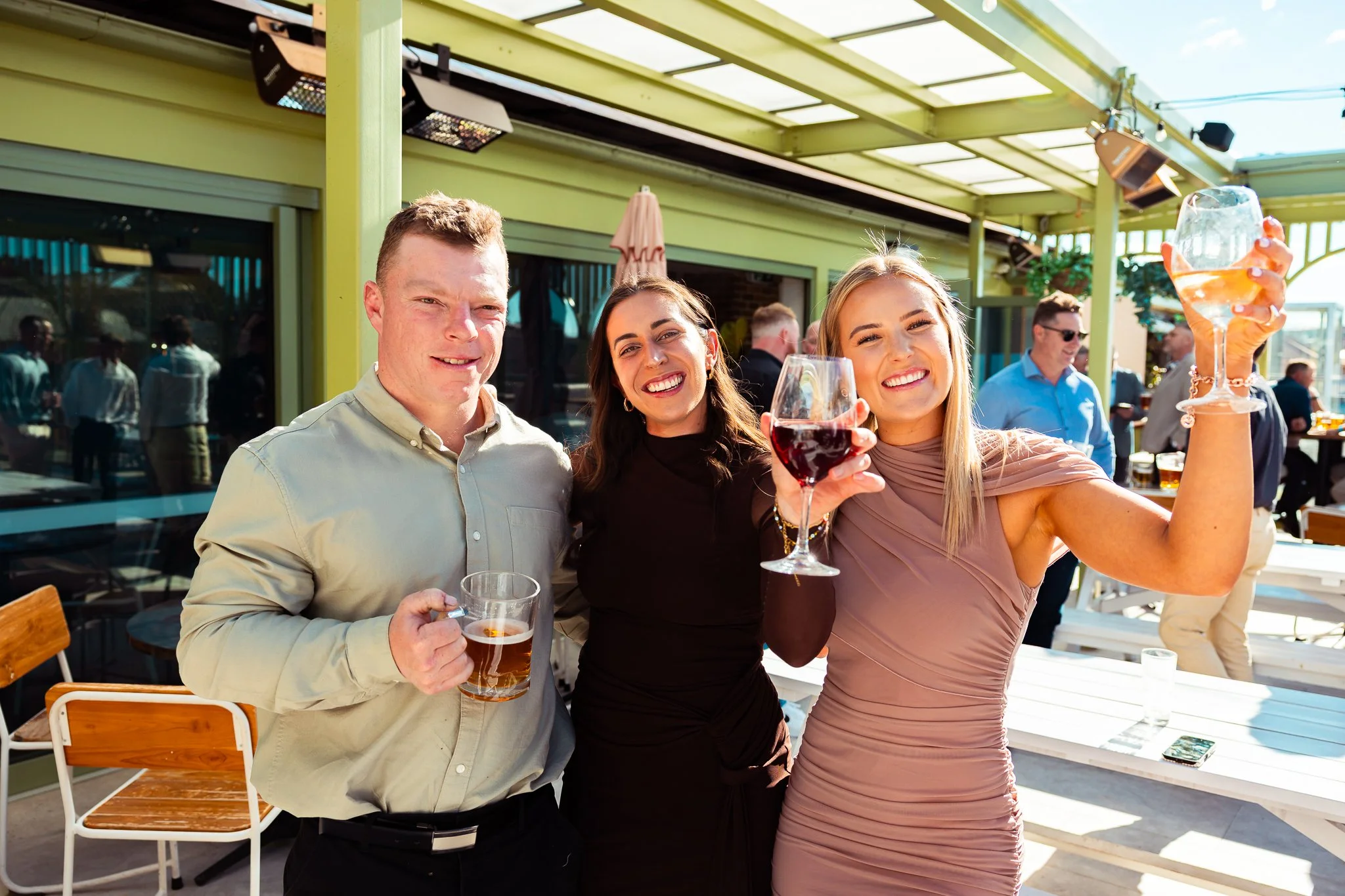 Three friends, two women and one man, enjoying drinks at an outdoor gathering on a sunny day. The woman on the right is holding a wine glass, the man has a beer mug, and the woman in the middle is smiling.
