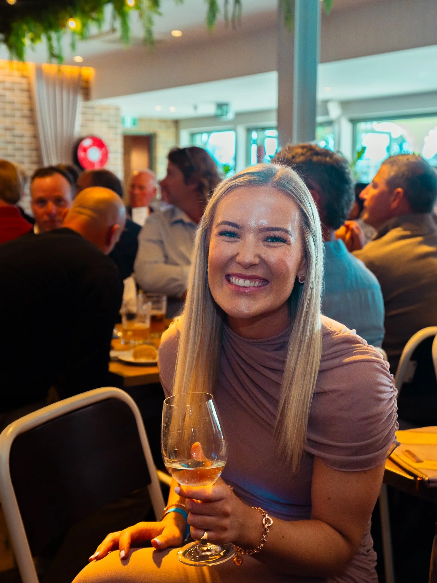 A woman with long blonde hair smiling and holding a glass of white wine at a social gathering in a bright, modern indoor setting.