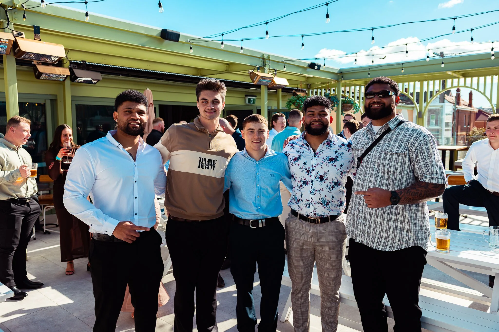 Group of five men smiling and posing together at an outdoor gathering on a sunny day, with other people in the background holding drinks and socializing.