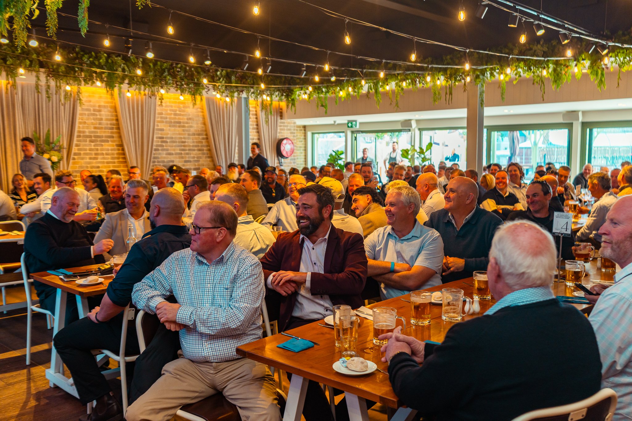 A large group of people, mostly men, sitting at long wooden tables enjoying a meal and drinks at a festive indoor event with warm lighting, string lights, and decorative greenery.