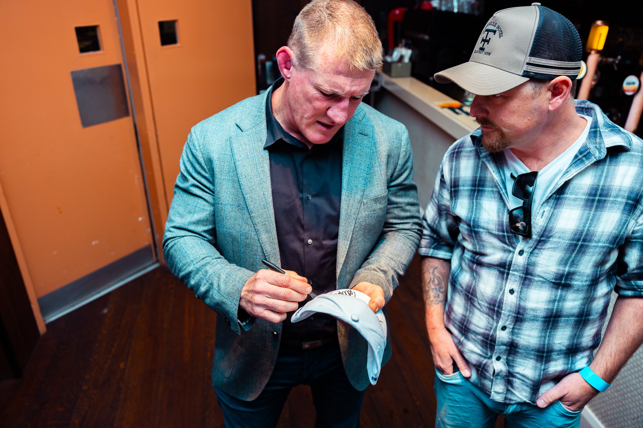 Two men are standing close together, looking at a sneaker that one of them is signing. One man is wearing a gray blazer and dark shirt, while the other is wearing a plaid shirt, sunglasses hanging from his collar, a cap, and has tattoos on his arm. T