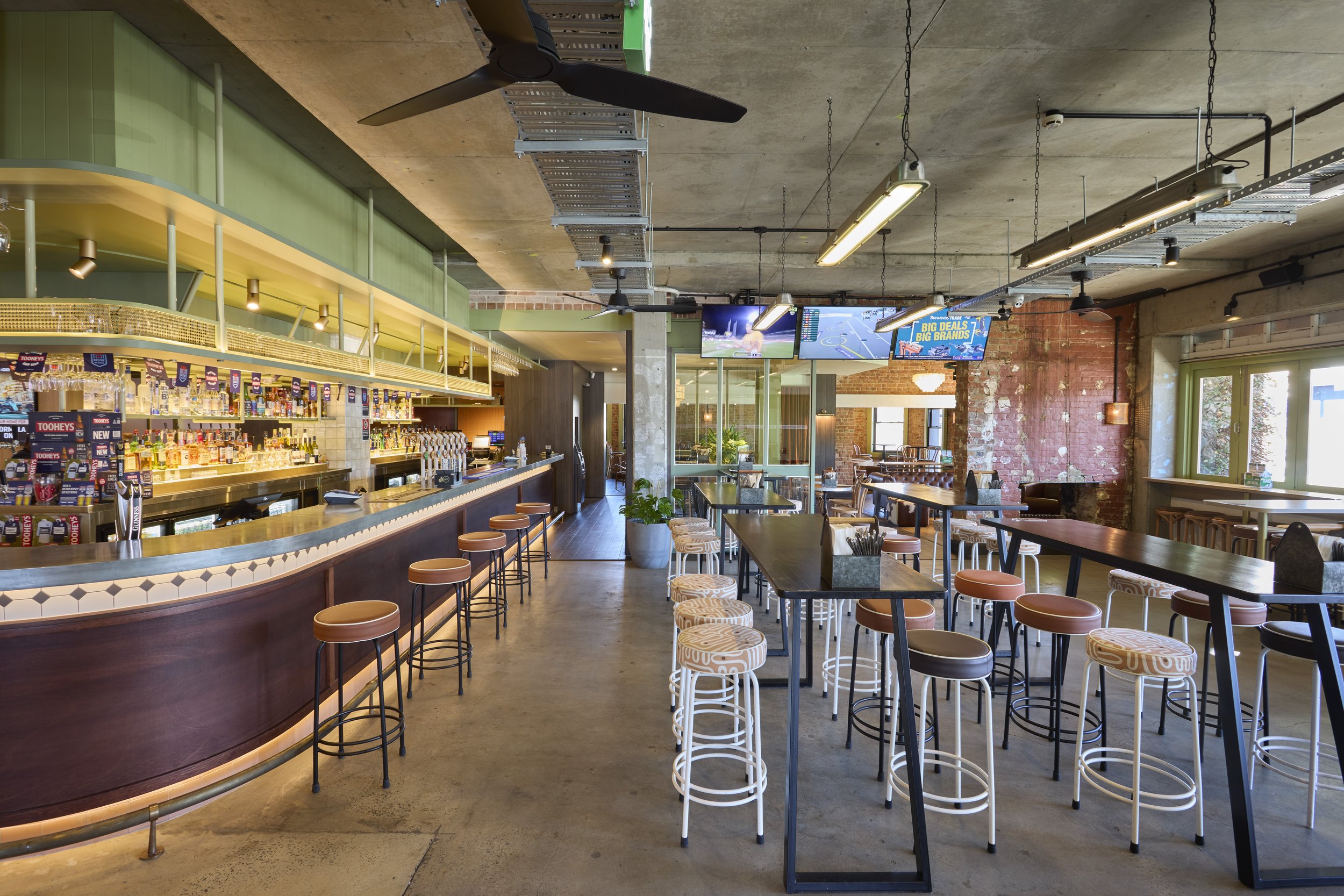 Interior of a modern bar or restaurant with a curved bar counter, high stools, multiple tables and chairs, exposed brick wall, ceiling-mounted TVs, and industrial lighting.