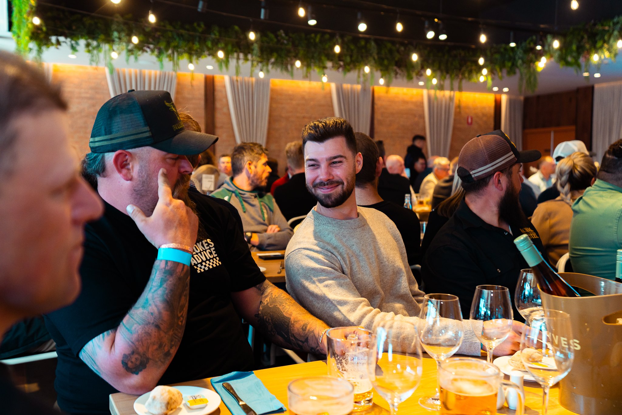 A group of people sitting at a long table in a restaurant or banquet hall, engaged in conversation, with food and drinks in front of them. The room has warm lighting with string lights and decorative greenery hanging from the ceiling.