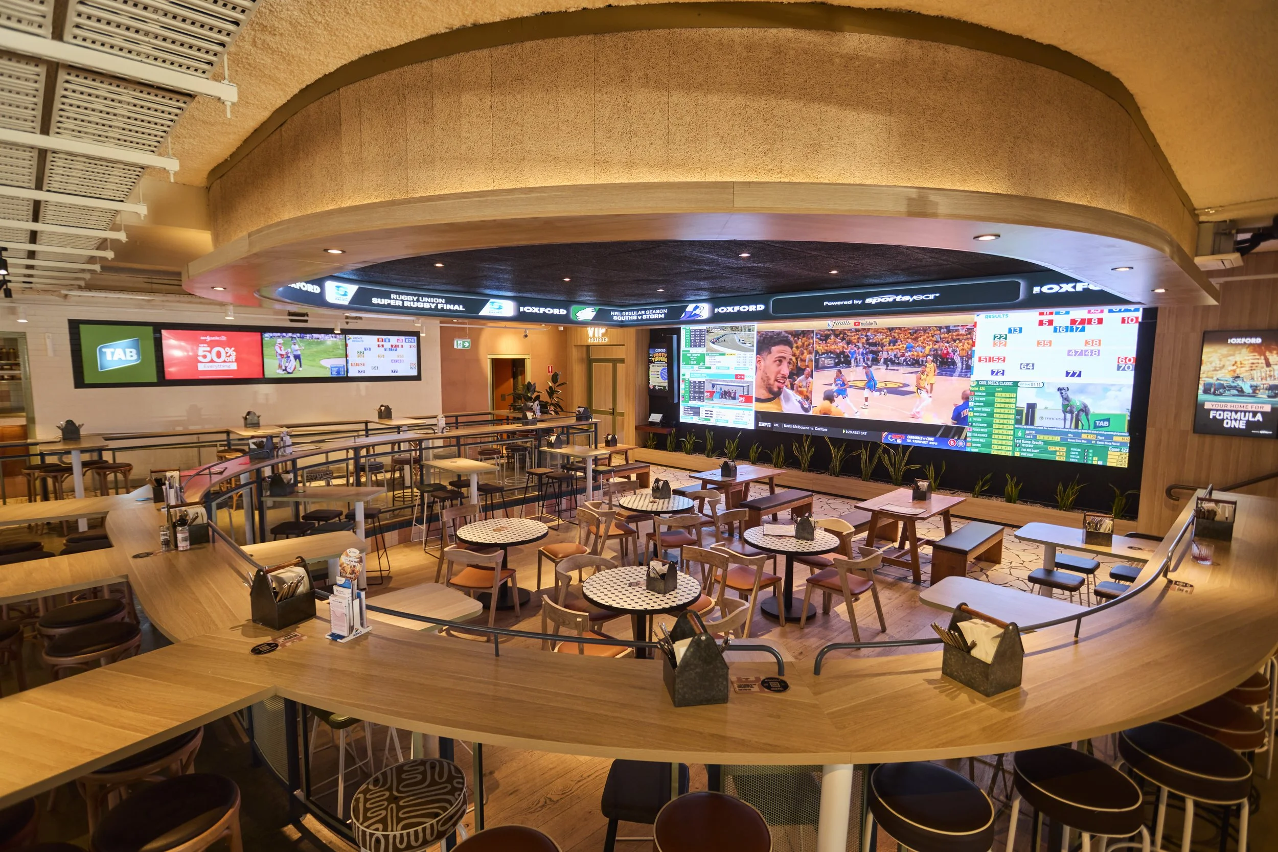 A sports bar or betting lounge with large screens displaying sports and betting odds. The room has tables and chairs, with some tables set for dining. The ceiling is curved with recessed lighting, and there is a wooden balcony railing overlooking the