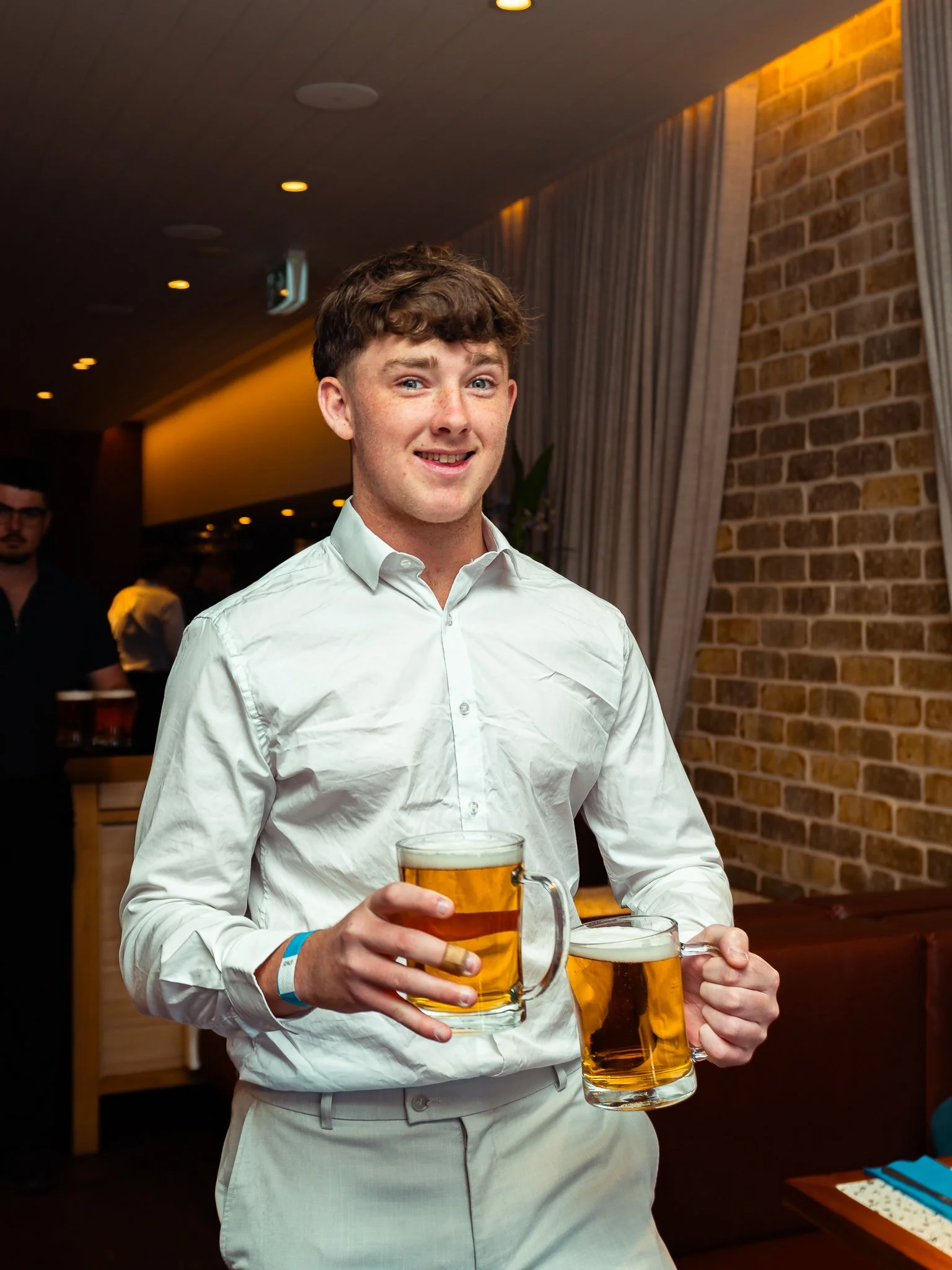 A young man in a white shirt holding two large mugs of beer. He is smiling and standing indoors, with a brick wall and curtains in the background.