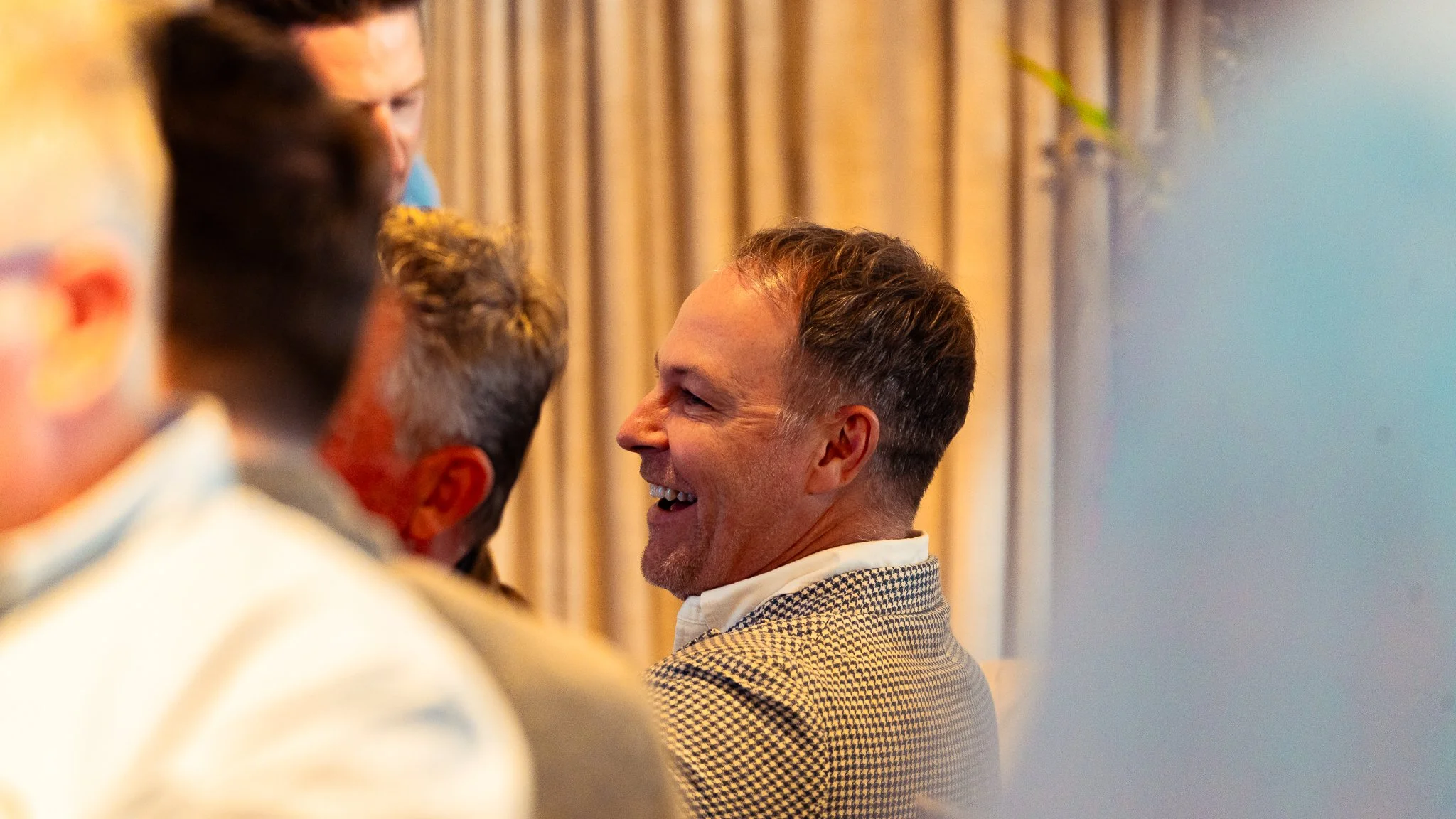 A group of laughing men engaged in conversation at an indoor event, with a beige curtain and a plant in the background.