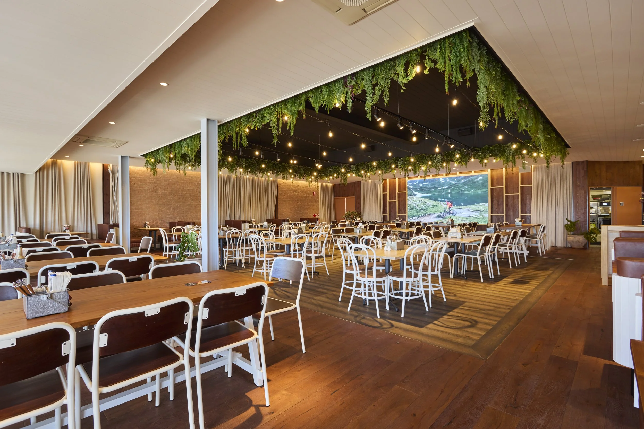 Inside a restaurant or event space with many tables and chairs, a large digital screen showing outdoor scenery, decorated hanging with greenery and string lights, and curtains on the walls.