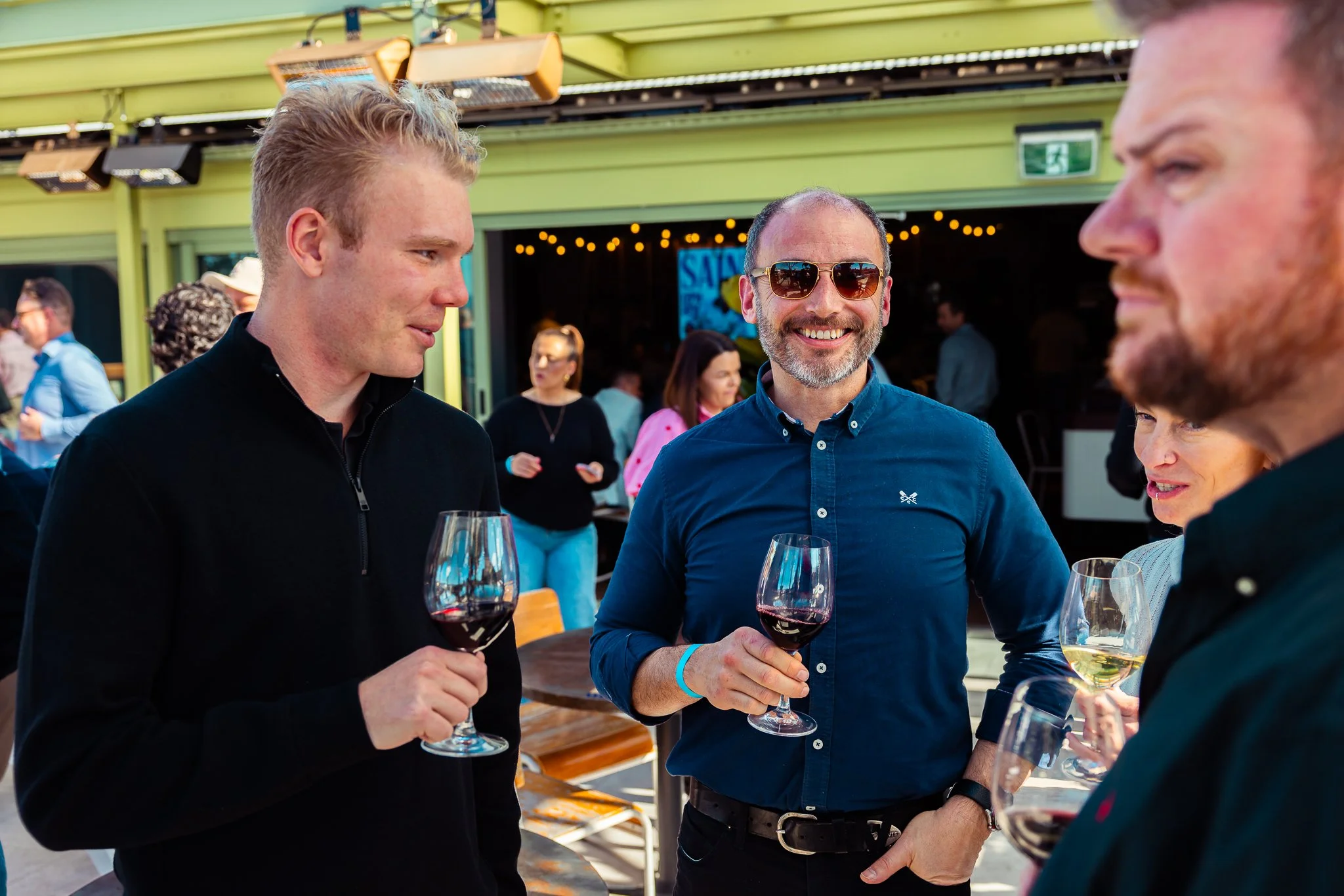 Three men holding glasses of red and white wine, engaging in conversation at an outdoor event with other people in the background.