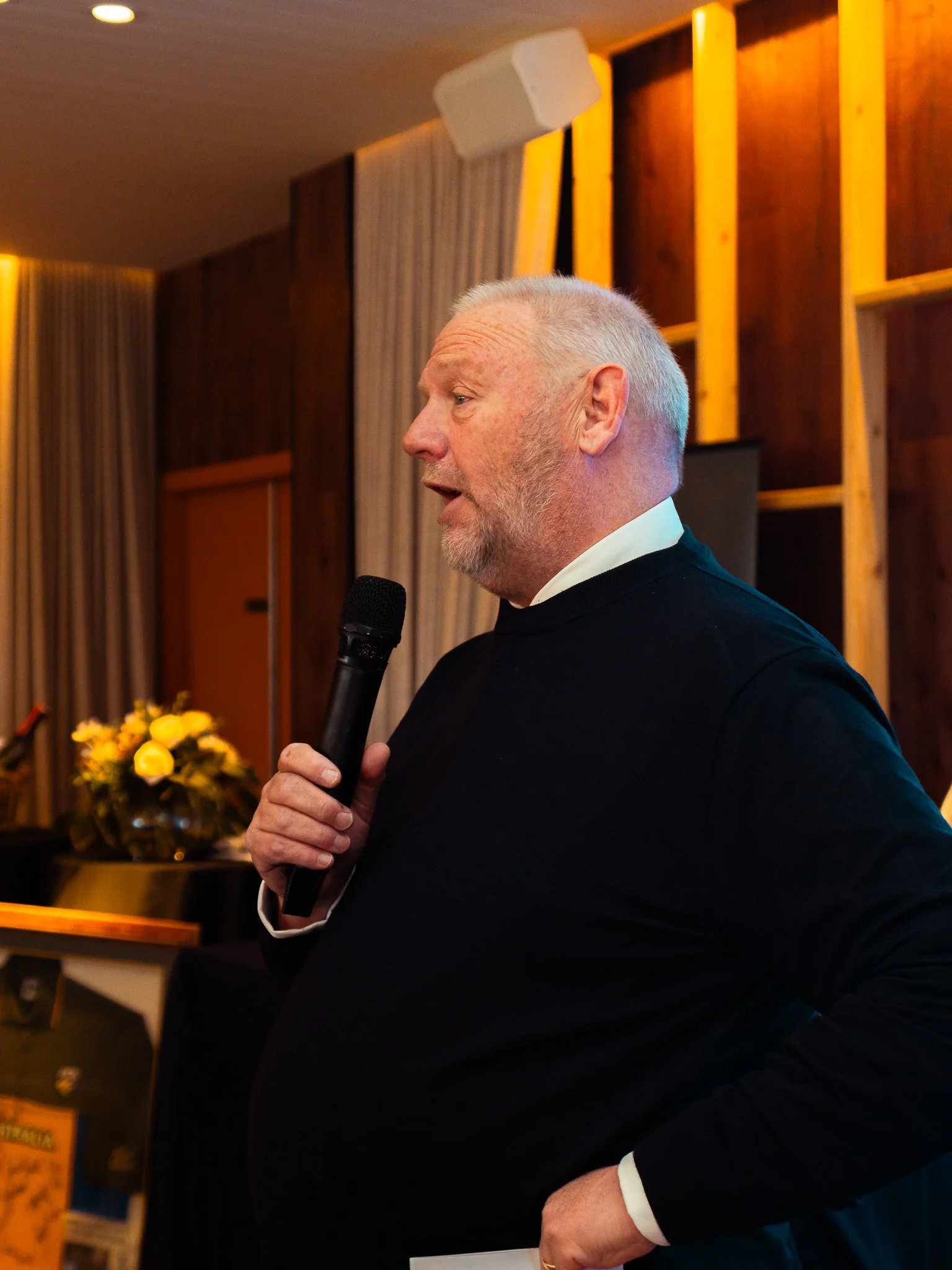 A man with gray hair and a beard holding a microphone, wearing a black suit with white cuffs, speaking at an indoor event.