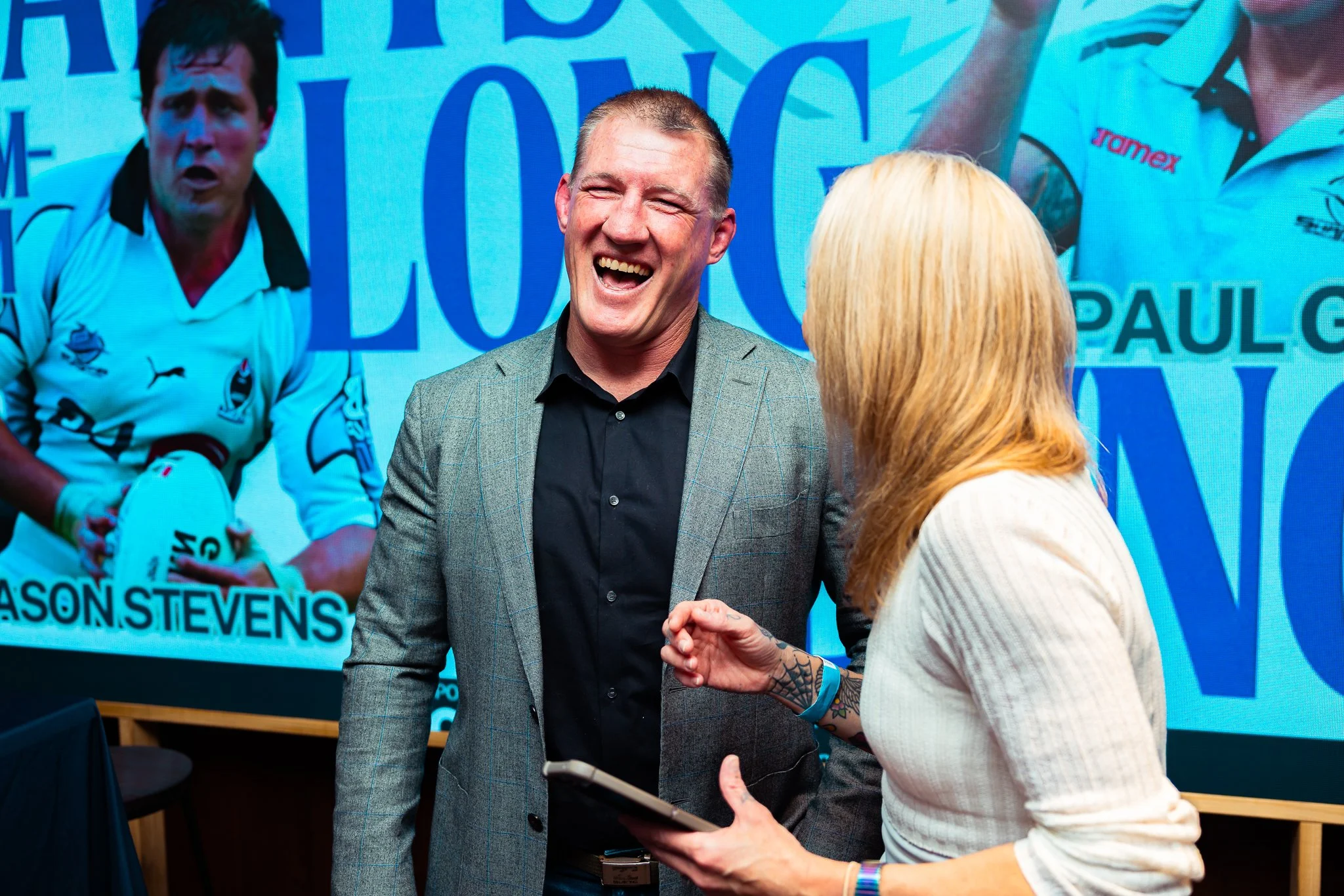 A man with short hair and a gray blazer is laughing and talking to a woman with blonde hair, who is holding a clipboard. In the background, there is a large screen displaying a cyclist in a white jersey and a blue background with text.