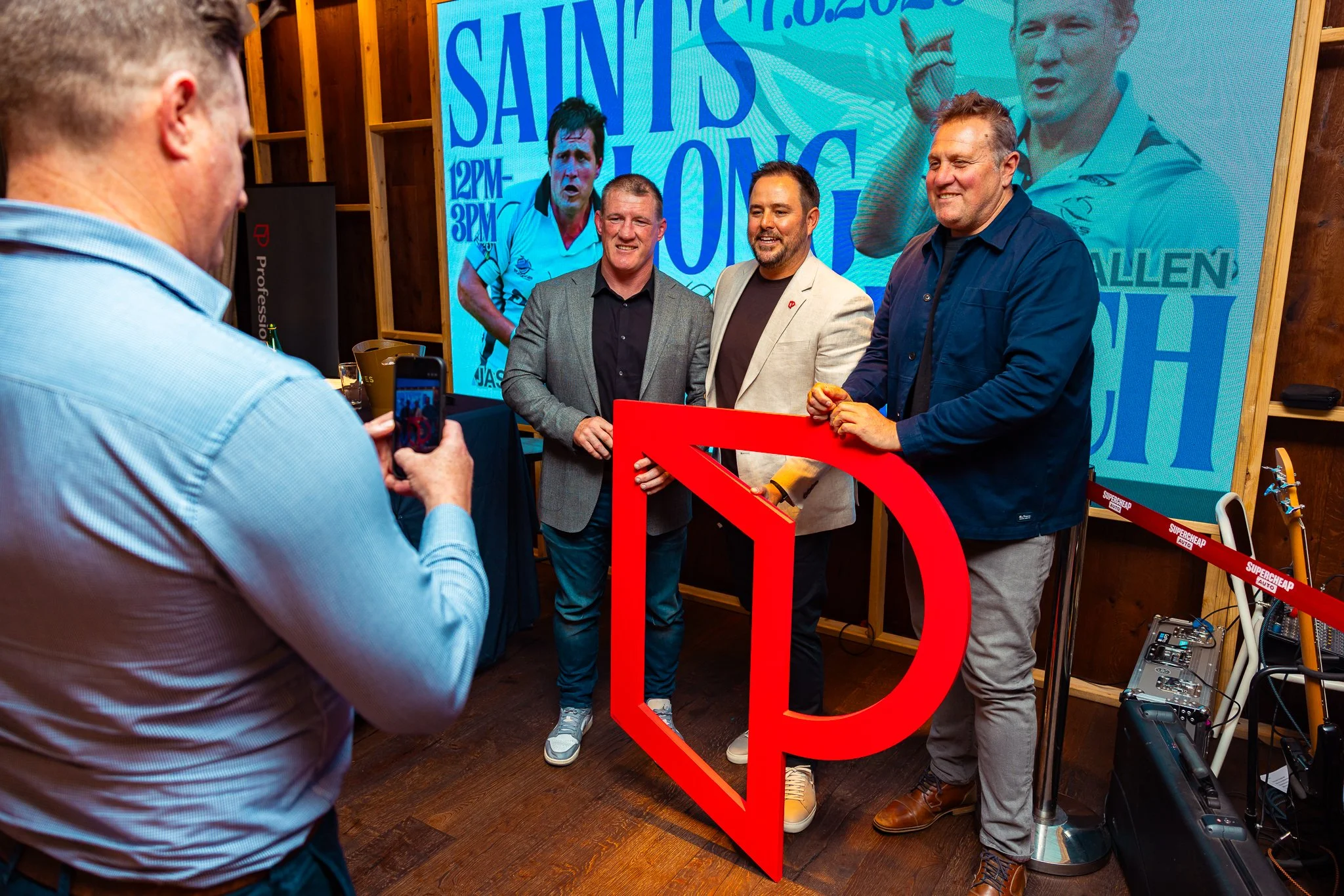 Four men are gathered at an event, with one taking a photo of the other three who are smiling and standing behind a large red letter D and a decorative frame. A large digital screen in the background displays blue text and images of athletes, indicat