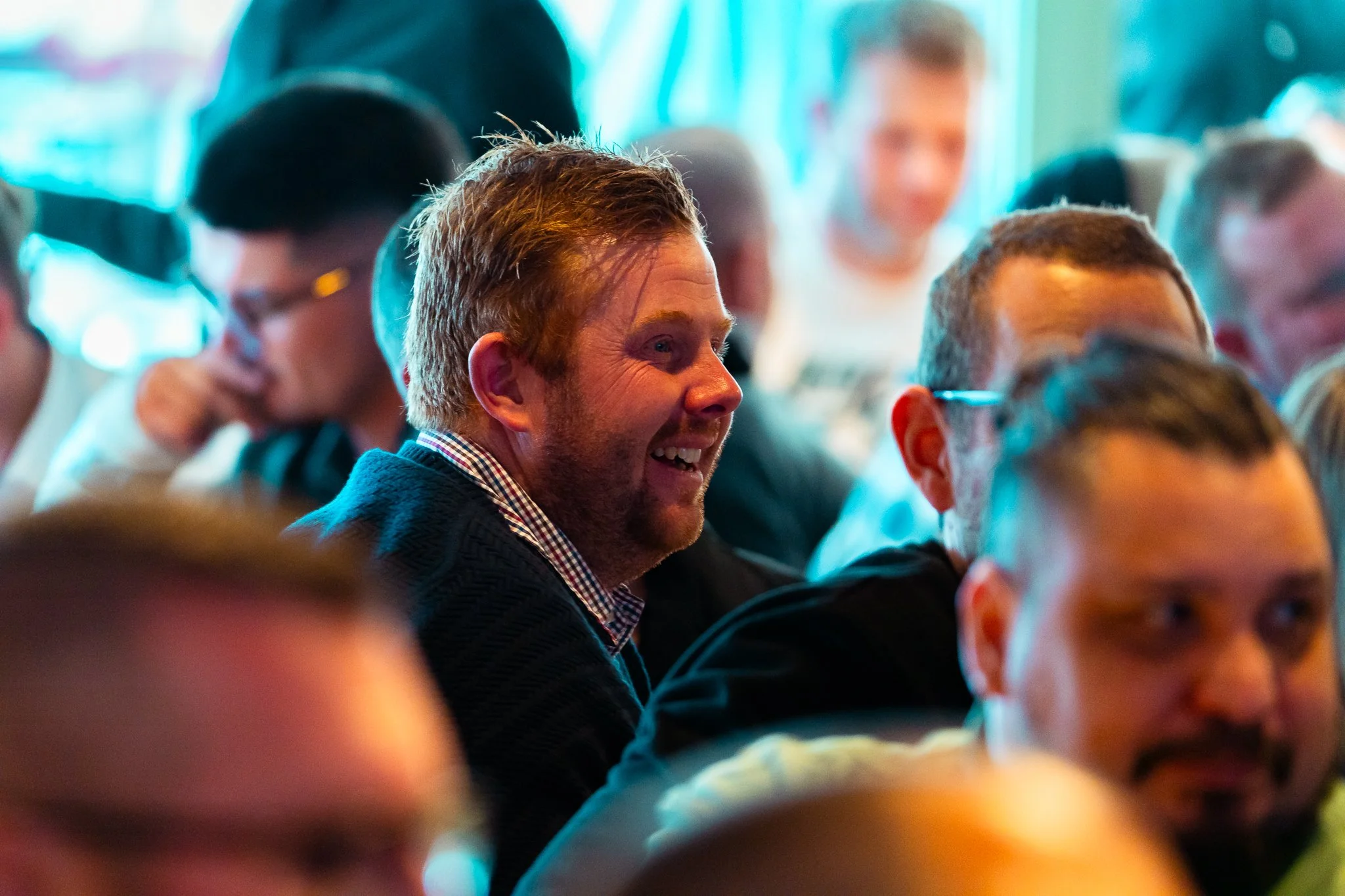 A group of people sitting at a conference or event, with one man smiling in focus in the foreground and other attendees in the background.
