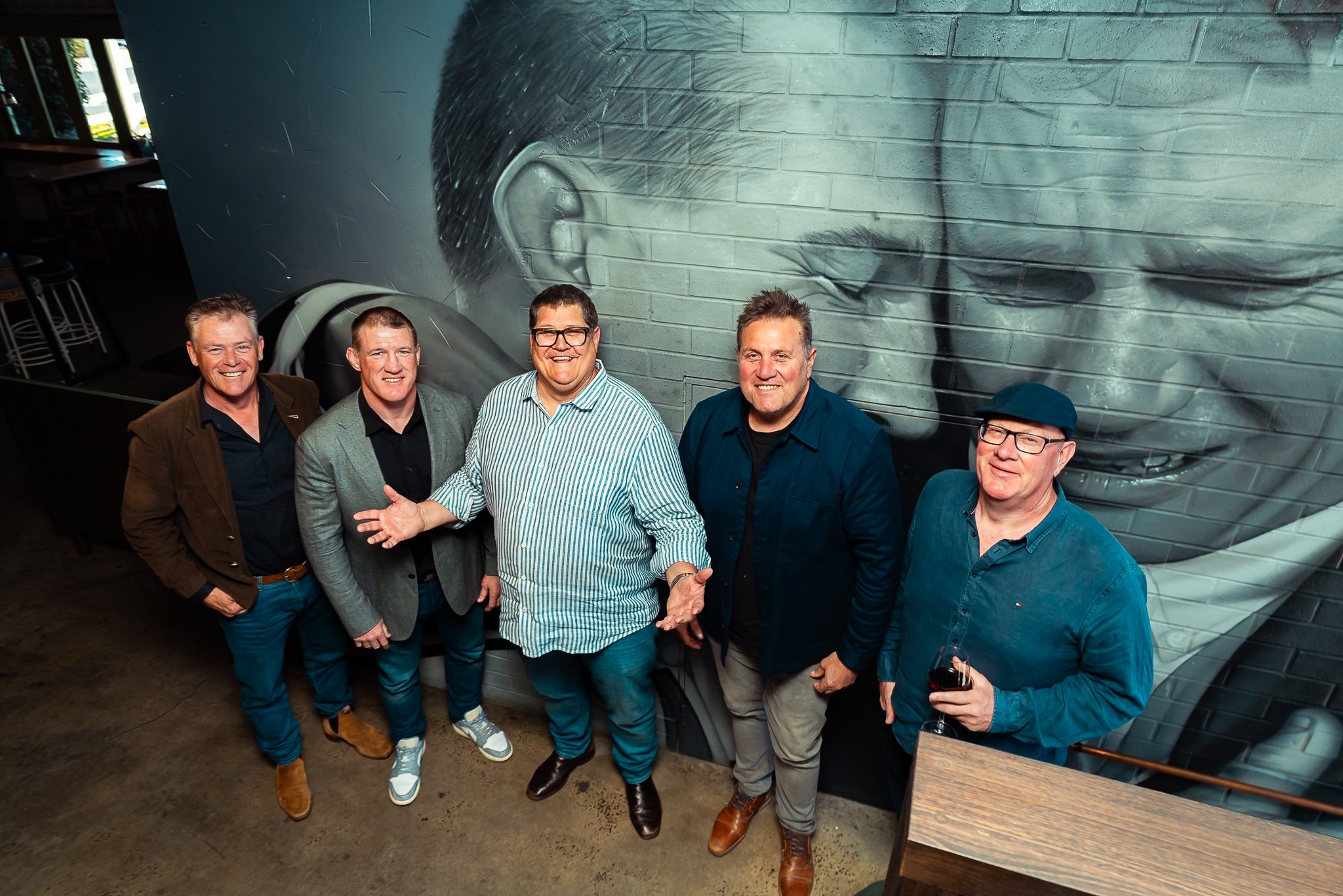 A group of five men standing together indoors, smiling for a photo in front of a large mural of a person's face on a brick wall.