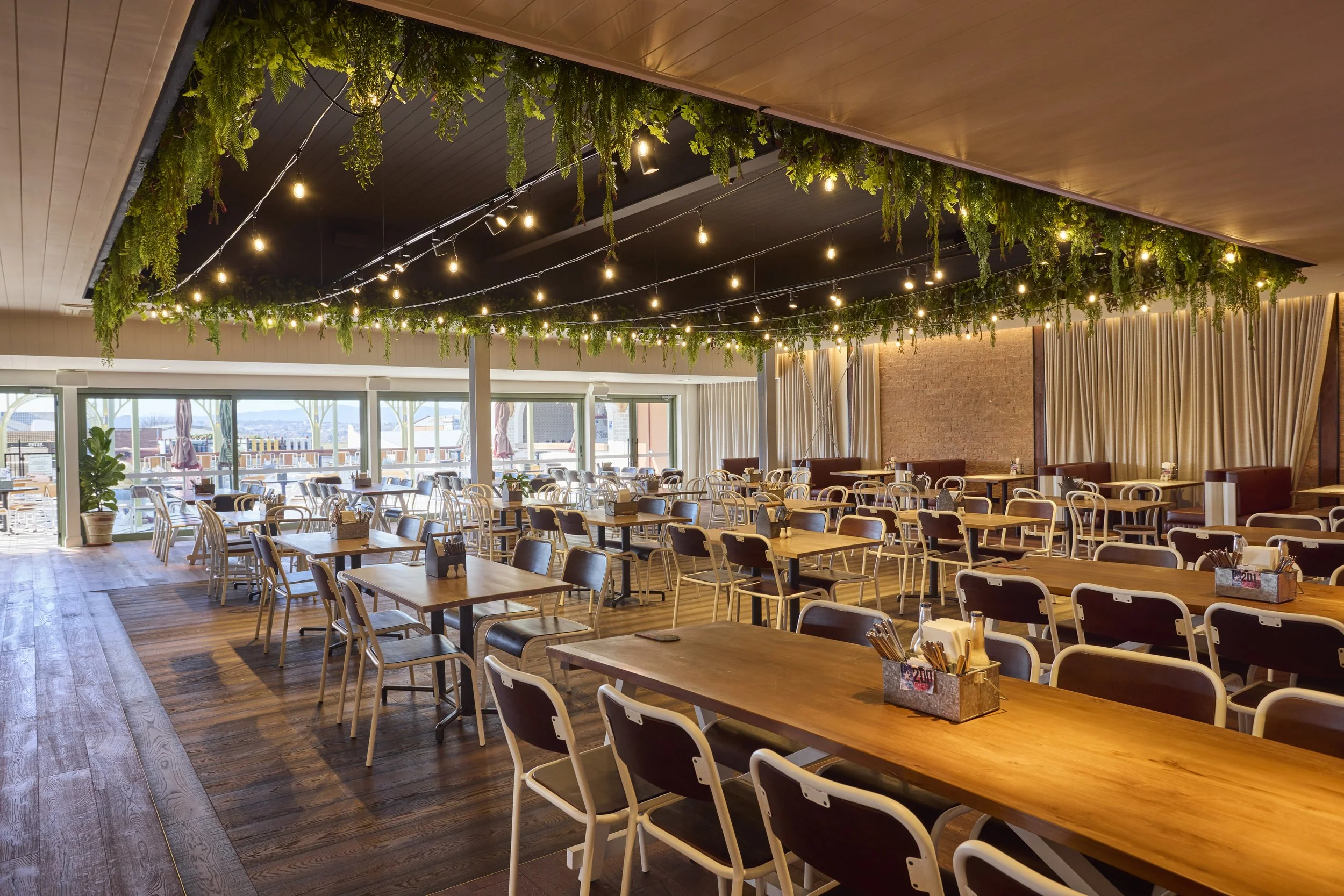Interior of a restaurant with wooden tables and chairs, hanging string lights, and a ceiling decorated with greenery. Large windows at the back reveal an outdoor patio area.