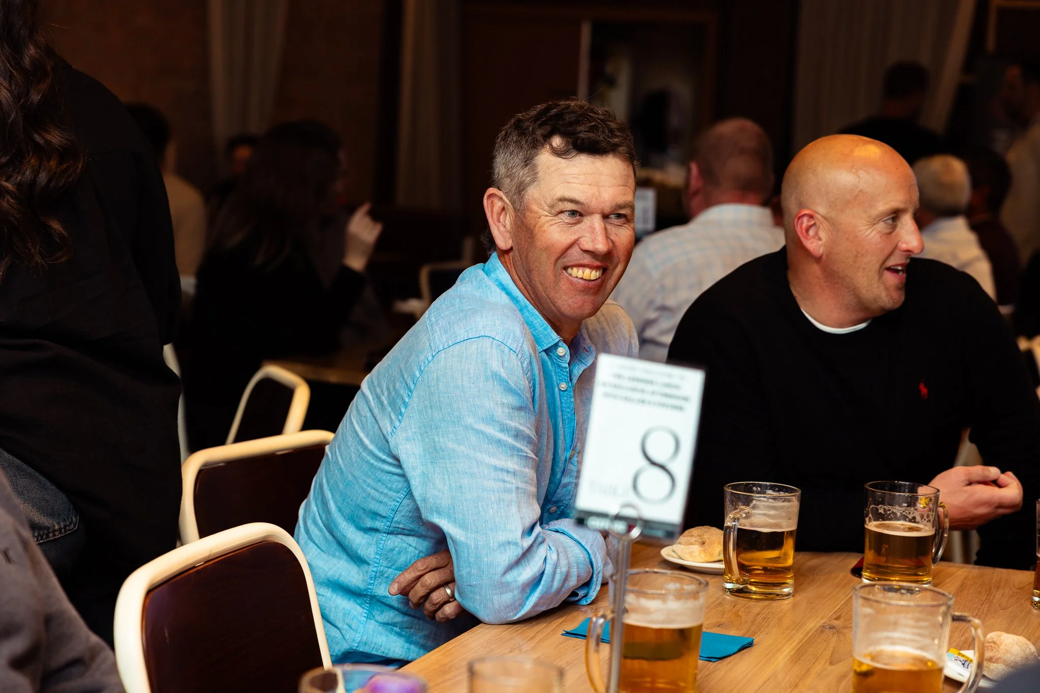 People seated at a table with drinks, engaged in conversation at an indoor social event.