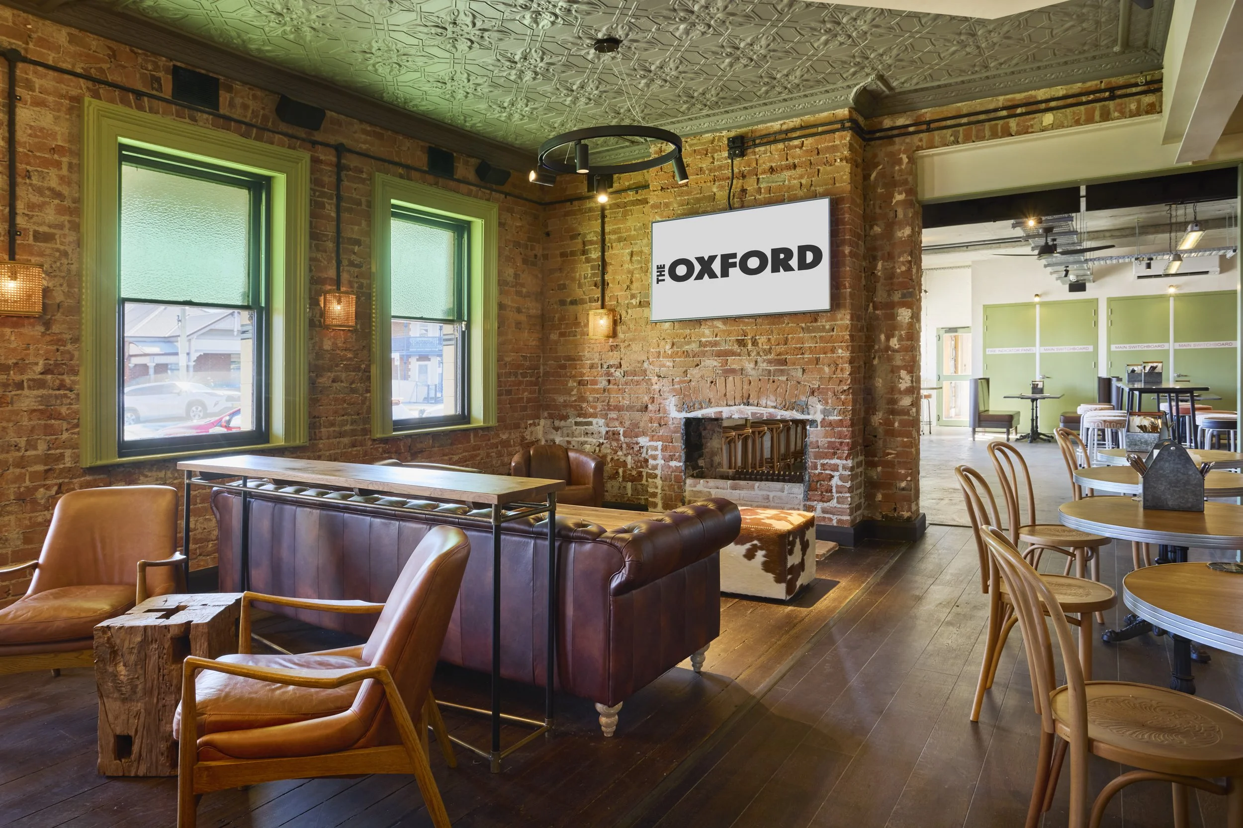 Interior of a cozy cafe or restaurant with exposed brick walls, large green-framed windows, leather and wooden chairs, a fireplace, and a flat screen TV displaying 'The Oxford' logo.